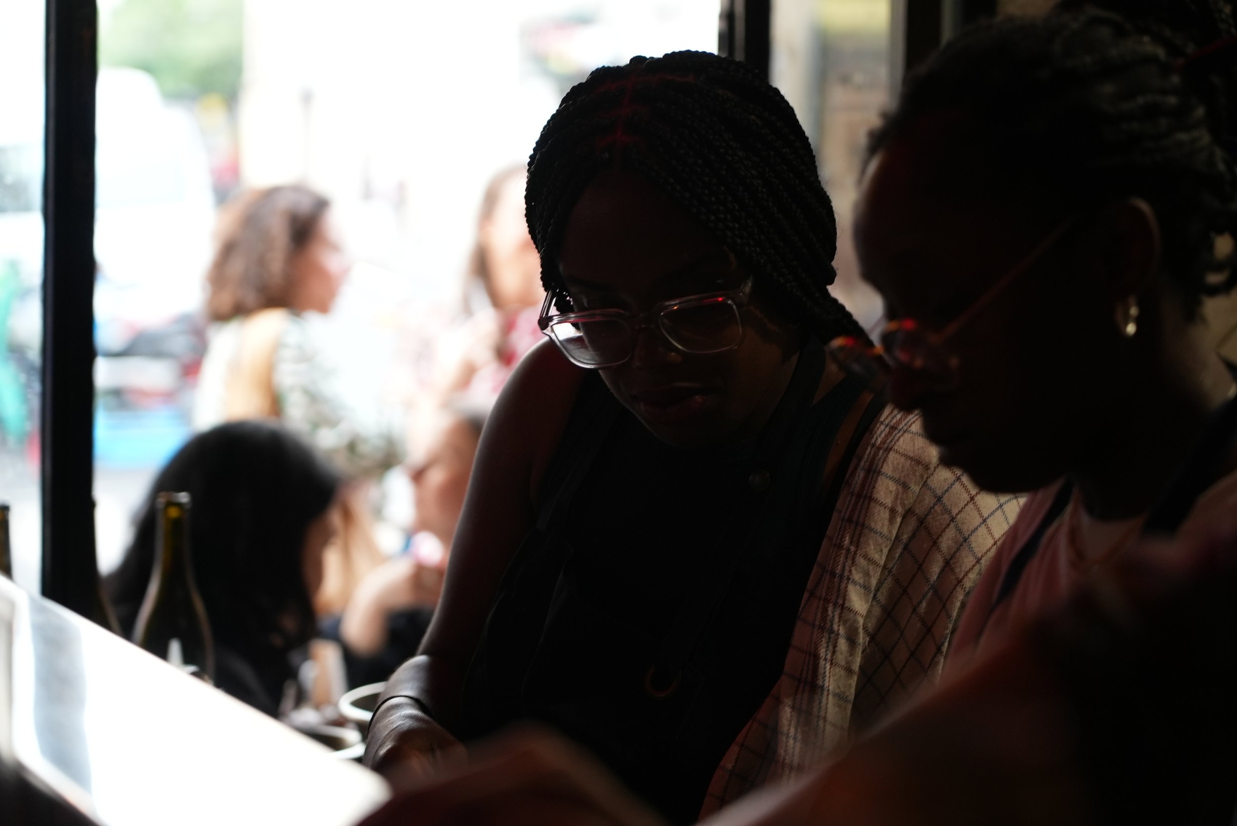 Two women with braided hair and glasses sitting at a bar or counter, with a group of women in the background outside a window.