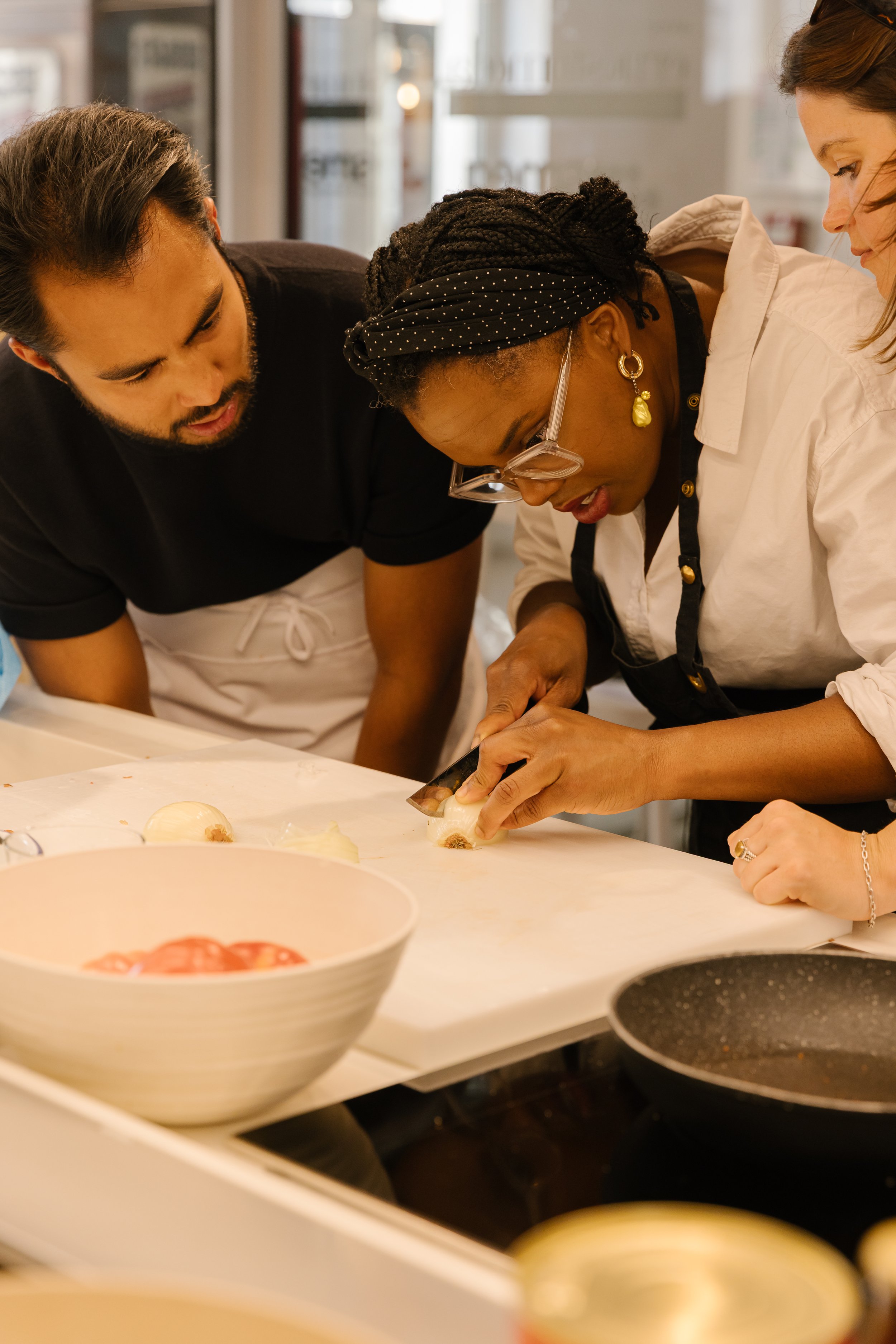 Three people are gathered around a kitchen counter, preparing food together. One person is wearing glasses and a headband, while the others are looking on and helping.