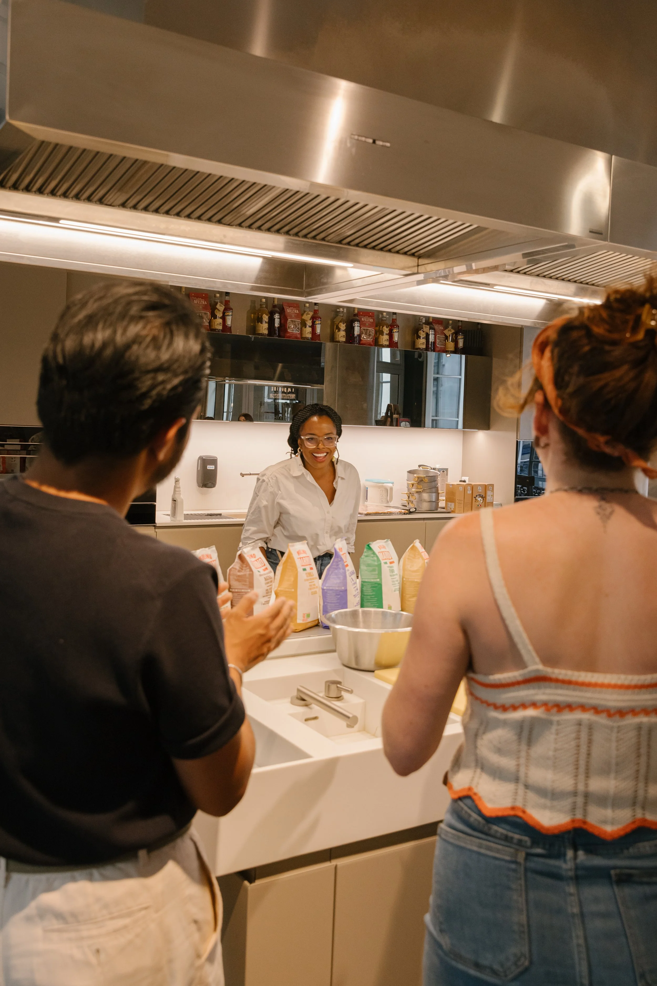 People gathered around a kitchen sink with a woman smiling in the background, kitchen counter with colorful snack bags and a large metal bowl.