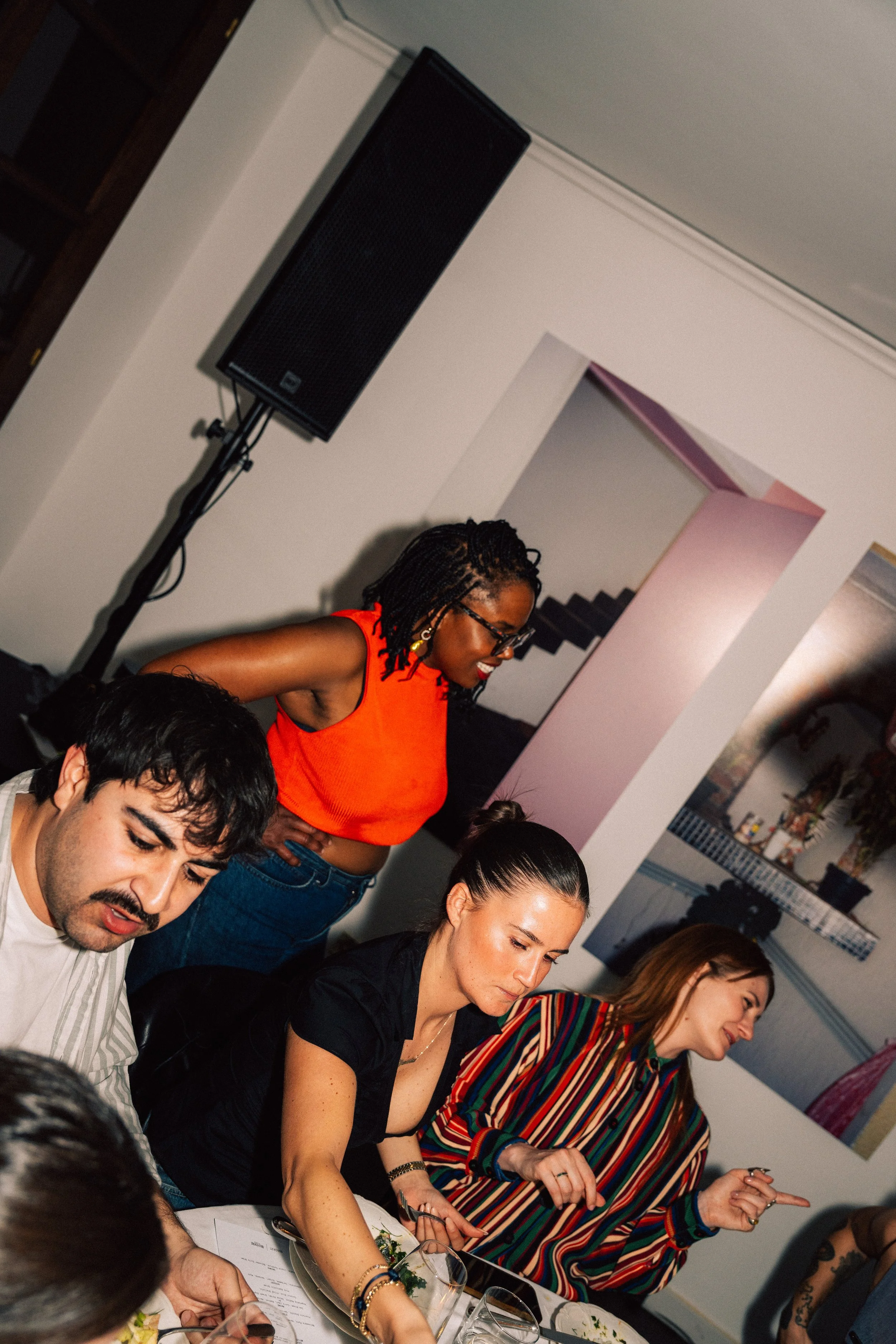 People sitting and standing at a table during a social gathering or dinner, with a woman in a colorful striped shirt pointing and talking.