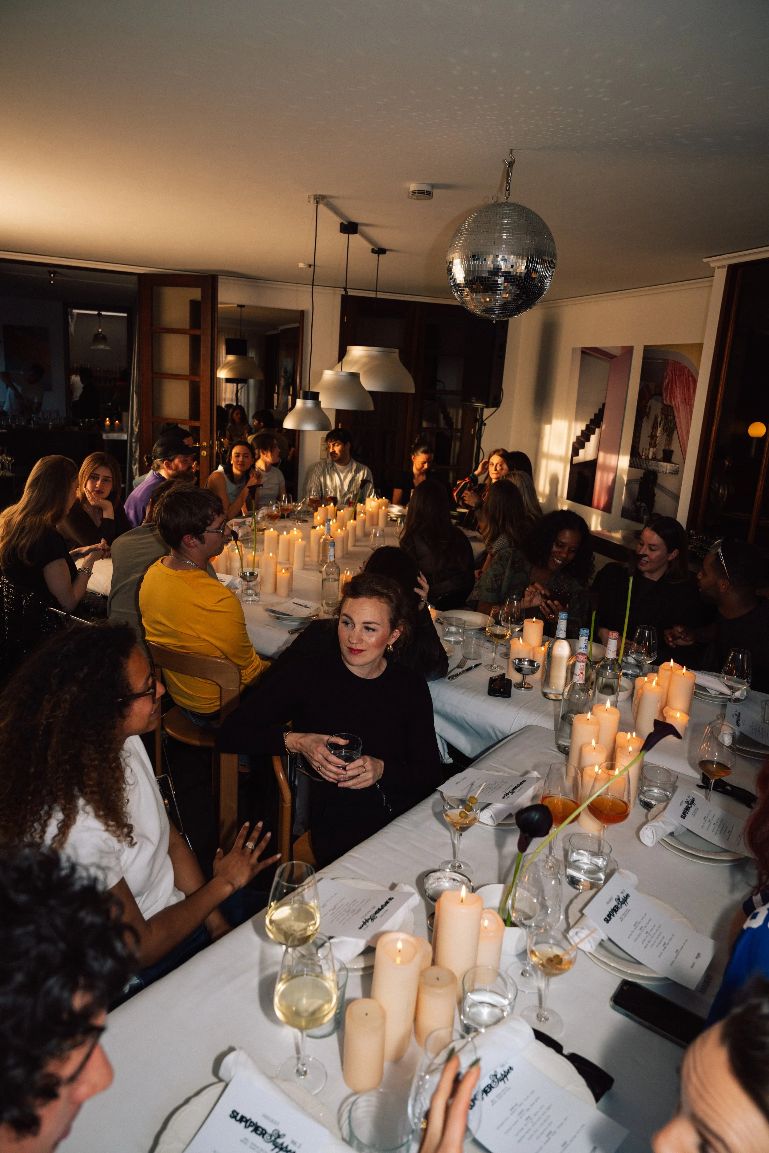 A large group of people gathered around a dinner table with lit candles, glasses of wine, and printed menus, in a warmly lit restaurant or private dining room