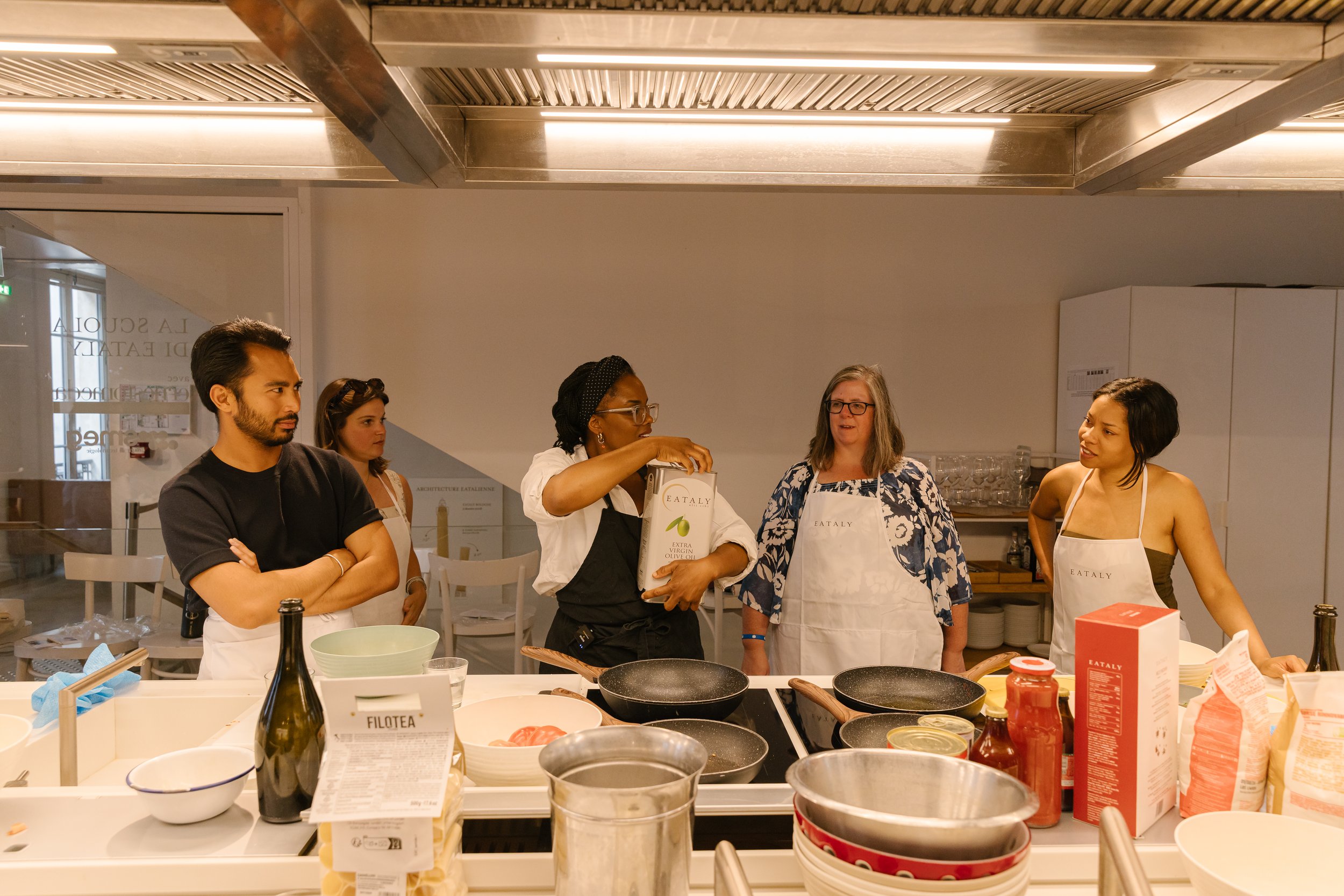 Group of five women in a kitchen, some wearing aprons labeled 'Eataly,' preparing for a cooking class or event. Various cooking ingredients and utensils are on the counter in front of them, including bottles, bowls, and packages.