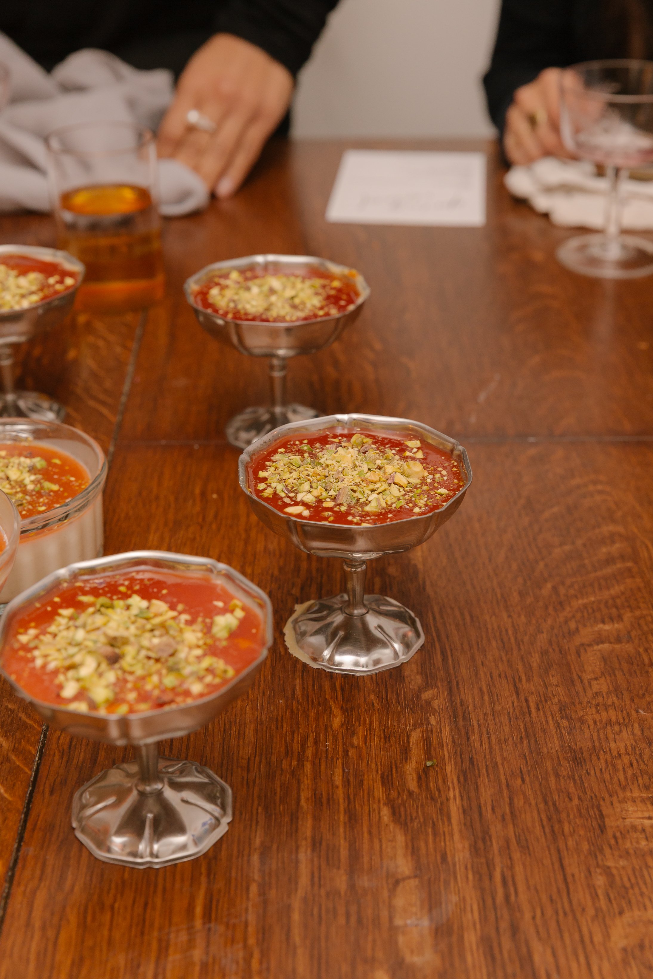 Several dessert cups filled with a red pudding topped with chopped nuts are arranged on a wooden table. People are seated around the table, with glasses and a paper note visible.