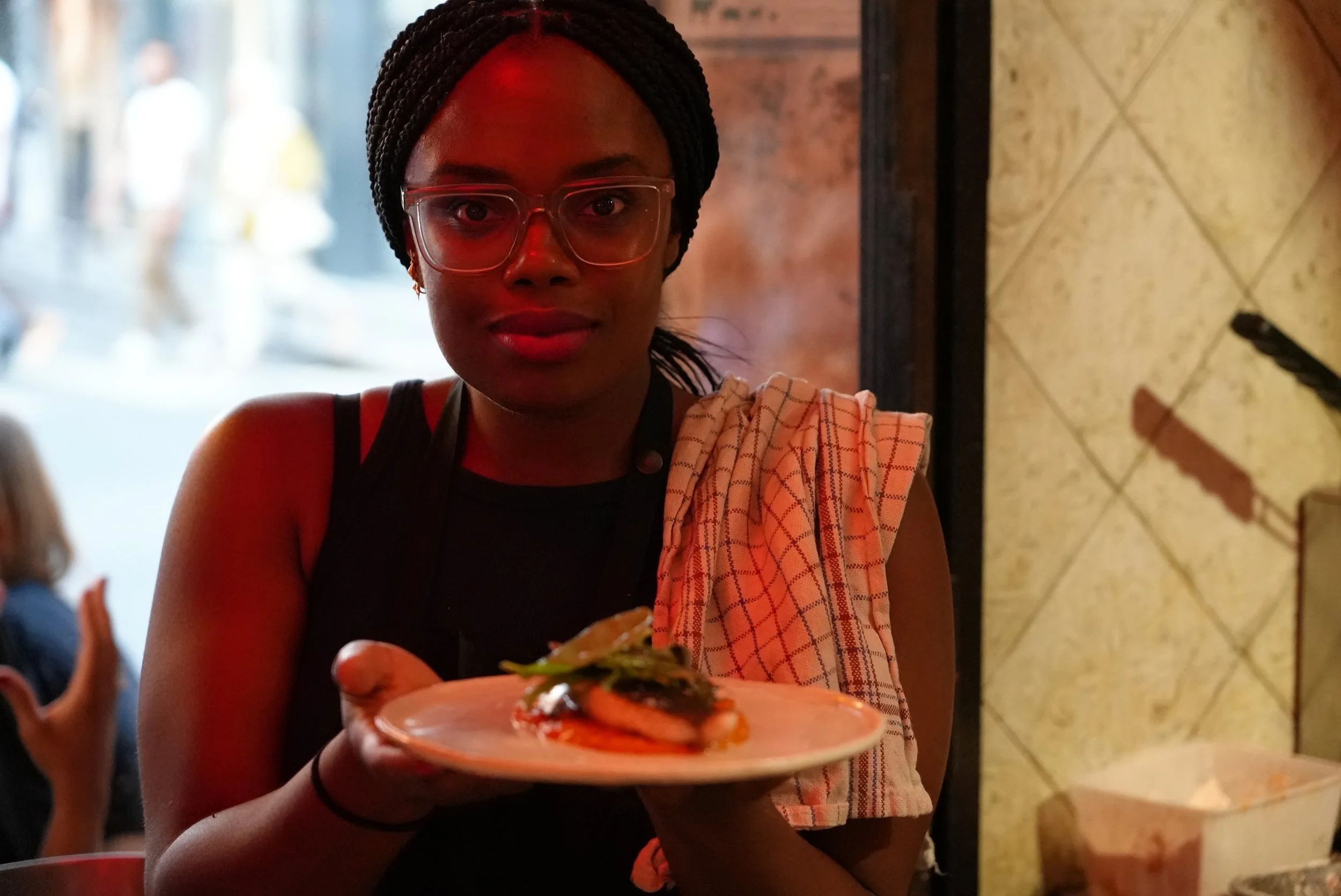 Woman wearing glasses and a black sleeveless top holding a plate with a fish dish, with a cloth draped over her shoulder, in a restaurant.