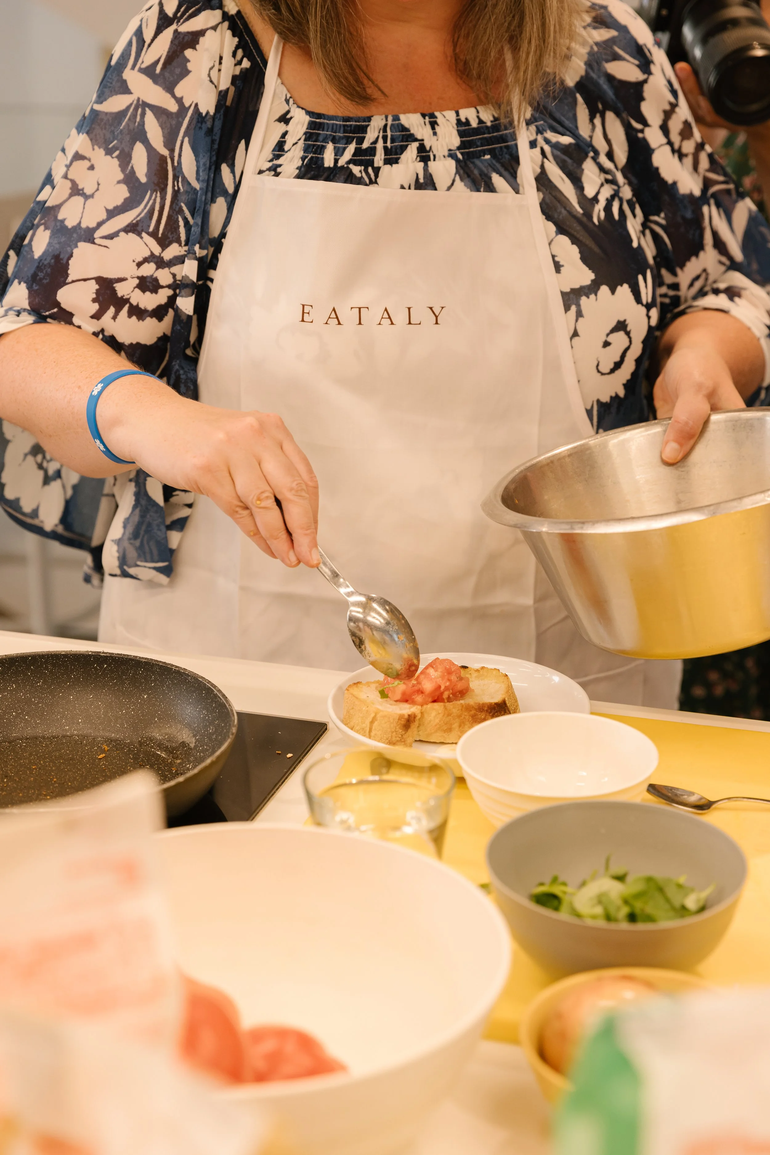 A woman wearing a white apron with 'EATALY' printed on it is preparing food in a kitchen, scooping toppings onto a slice of bread. There are various bowls and cooking utensils on the counter.