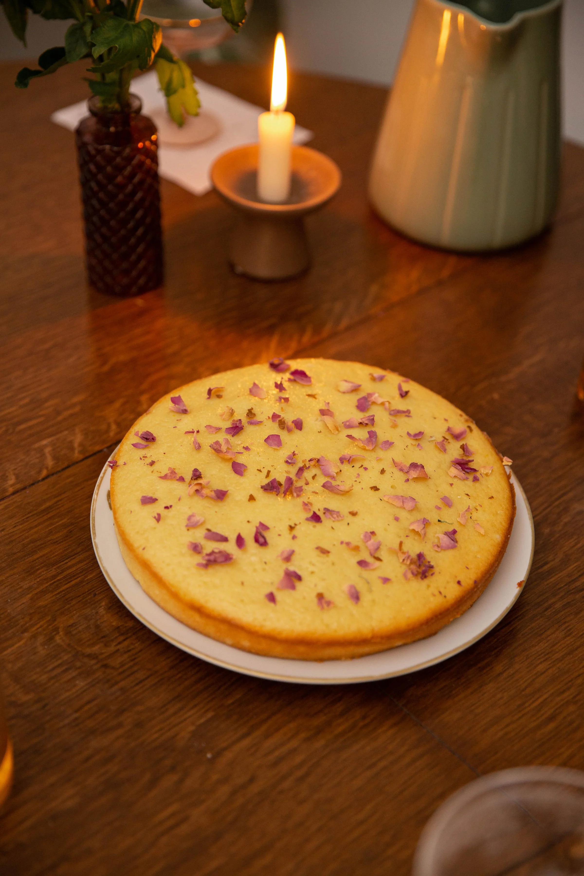 A yellow cake with pink rose petals on top on a white plate, placed on a wooden table. In the background, there is a lit candle in a holder, a brown vase with green leafy plant, a mug, and a white napkin.