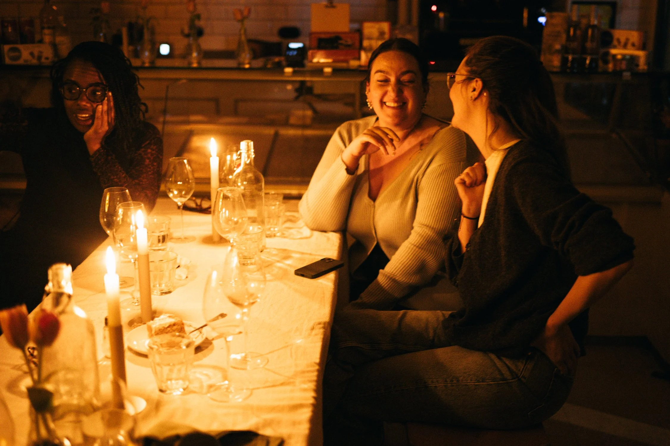 Three women sitting around a candlelit dinner table, laughing and talking with each other at a cozy, dimly lit restaurant.