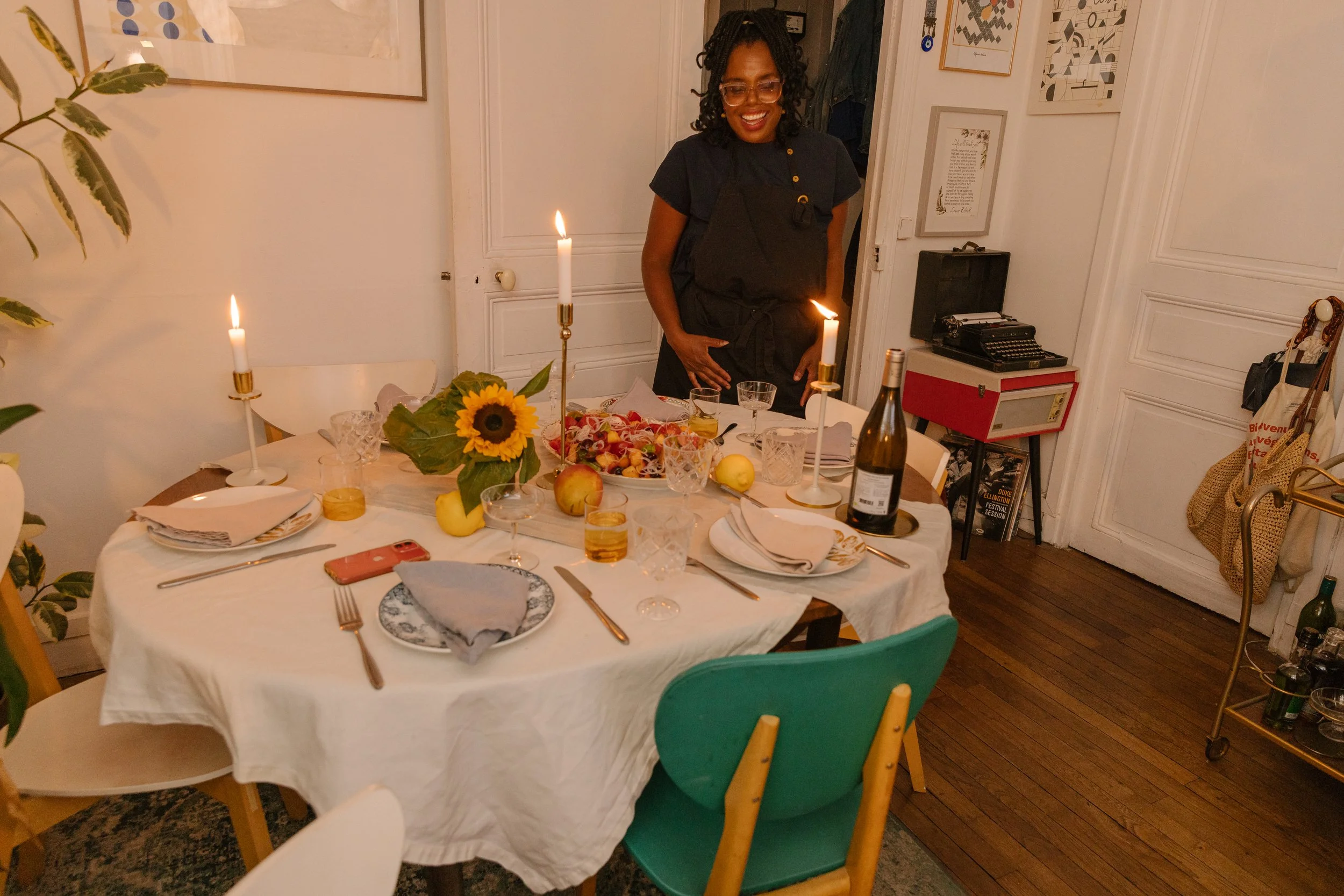 Woman smiling at a dining table set for a celebration with candles, flowers, and food. The room has white walls decorated with framed artwork, and there are chairs of various colors around the table.