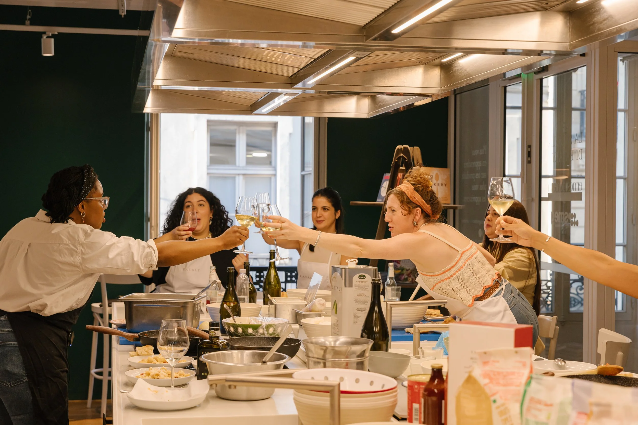 A group of women at a cooking class raising their glasses in a toast. The kitchen counter in front of them is filled with bowls, bottles, and ingredients.