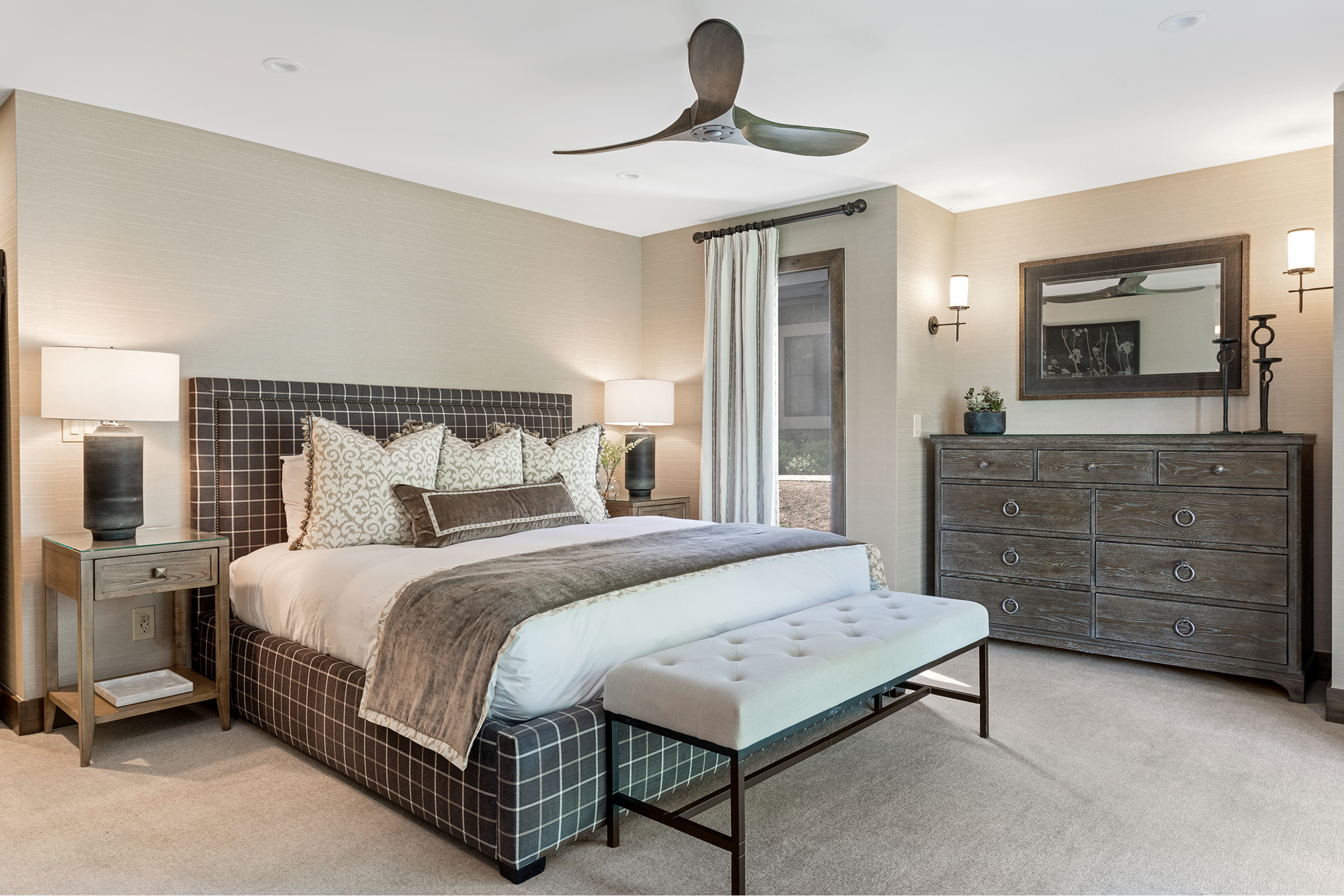 A bedroom with a large bed, a bench at the foot, two nightstands with lamps, a dresser with a mirror, a wall-mounted lamp, and a ceiling fan.