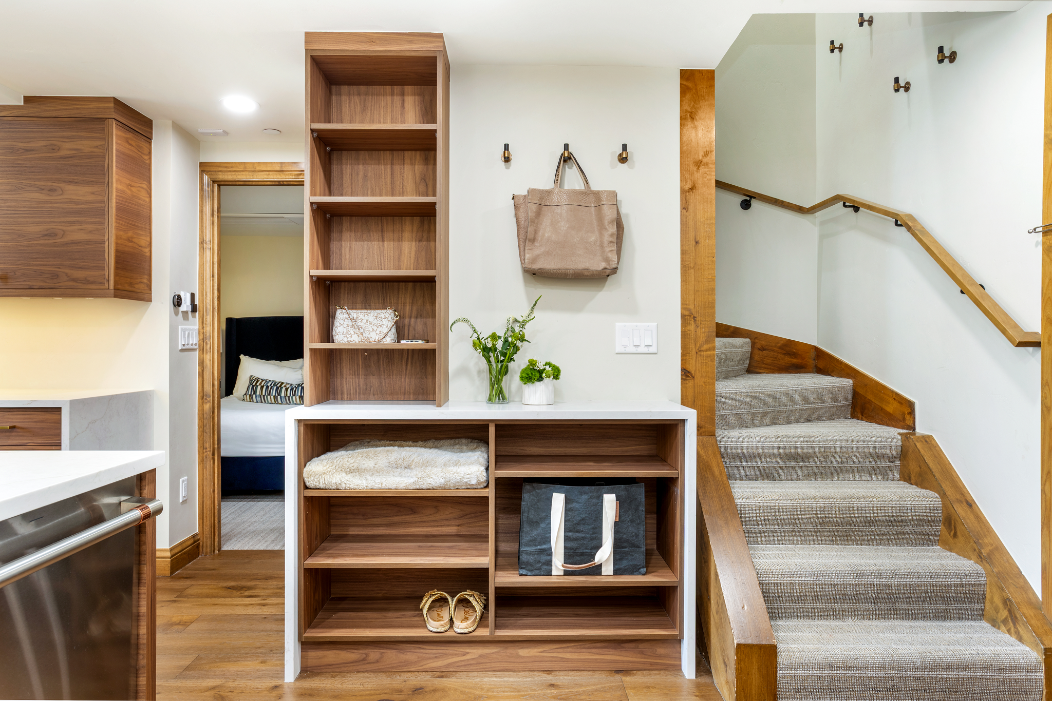 Interior of a house with wooden stairs, built-in wooden shelving, and a doorway leading to a bedroom. The wall has hooks with bags and floral arrangements underneath. Part of kitchen and laundry appliances are visible.