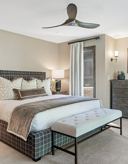 Modern bedroom with a plaid upholstered bed, a white bench at the foot, beige walls, a window with white curtains, a chest of drawers, and a ceiling fan