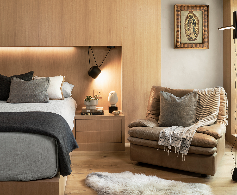 A cozy bedroom corner featuring a bed with gray and white bedding and multiple pillows, a floating nightstand with a plant, books, and a small lamp, a leather armchair with a throw blanket and pillows, and wall art depicting Our Lady of Guadalupe. There is a white fluffy rug on the light wooden floor.