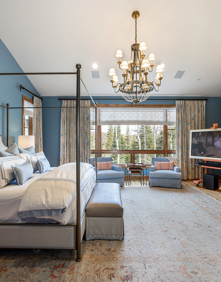 Living room with blue walls, four-poster bed, chandelier, seating area with two armchairs, a large window with curtains, and a TV.