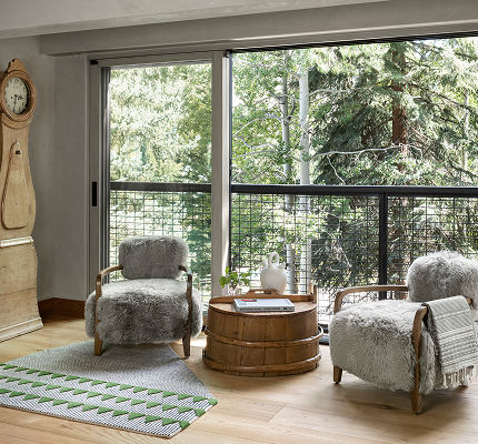 Living room with two armchairs with fuzzy gray cushions, a round wooden coffee table with a white teapot and magazines, a patterned rug, and large glass sliding doors leading to a balcony with trees outside.