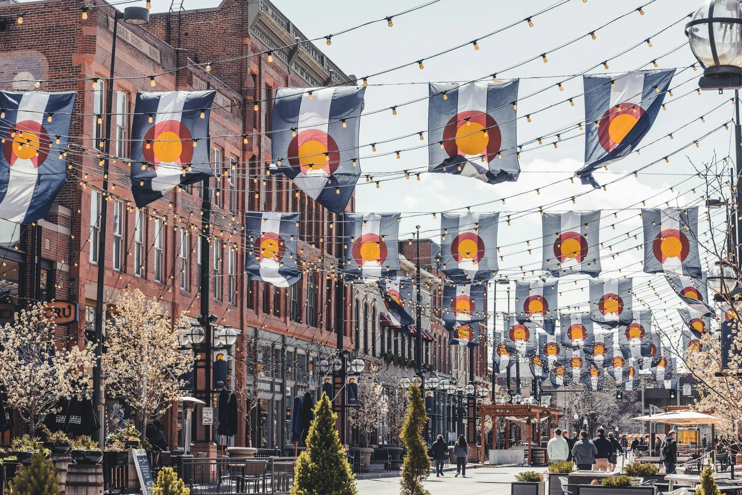 Larimer Square's main street decorated with Colorado flags and string lights, with people walking and blooming trees lining the sidewalk.