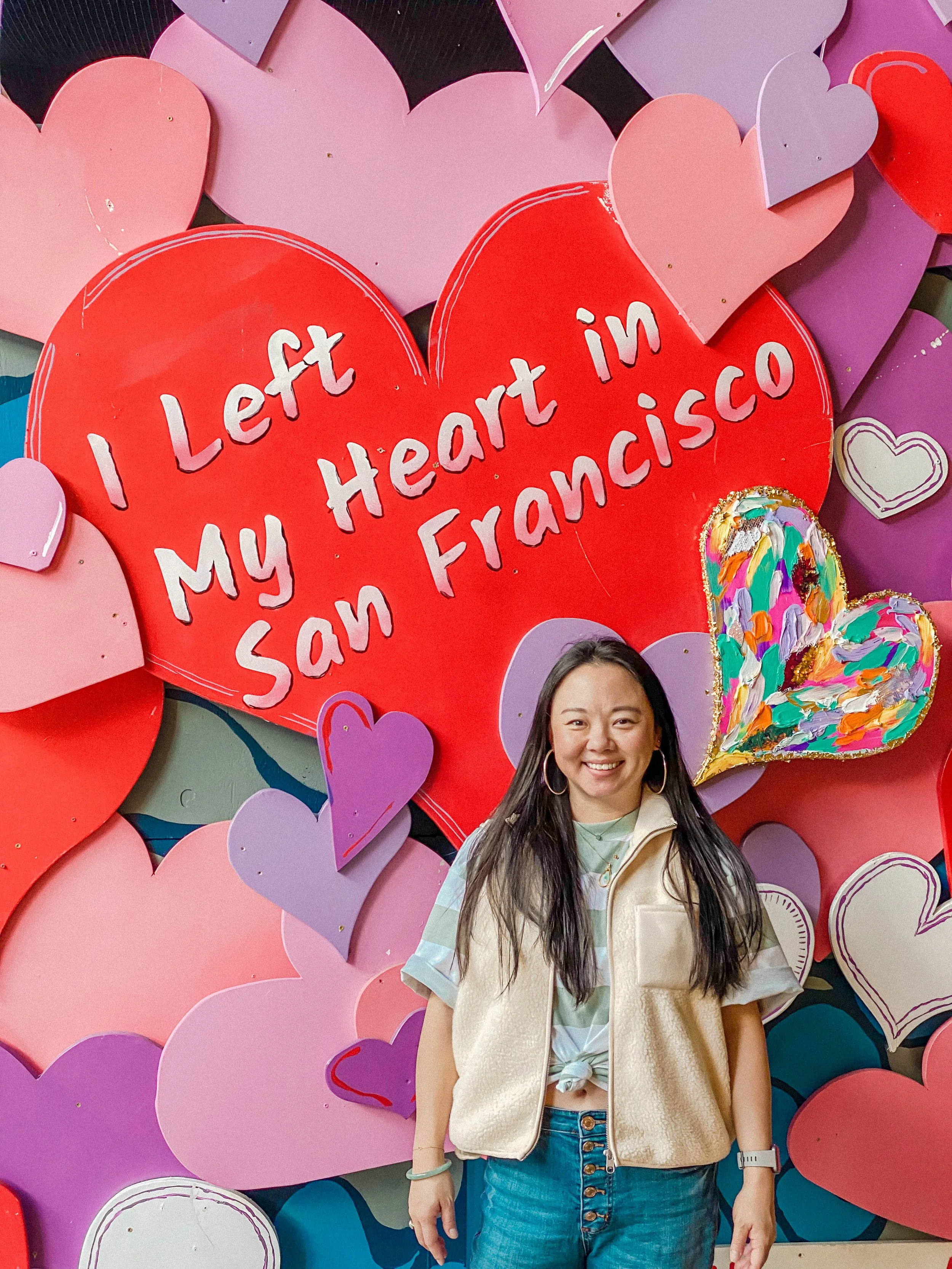A woman standing in front of a wall decorated with pink, purple, and red heart shapes; a large red heart in the center has the text 'I Left My Heart in San Francisco' written on it; the woman is smiling and wearing a cream-colored vest, a green and white striped shirt, and denim jeans.