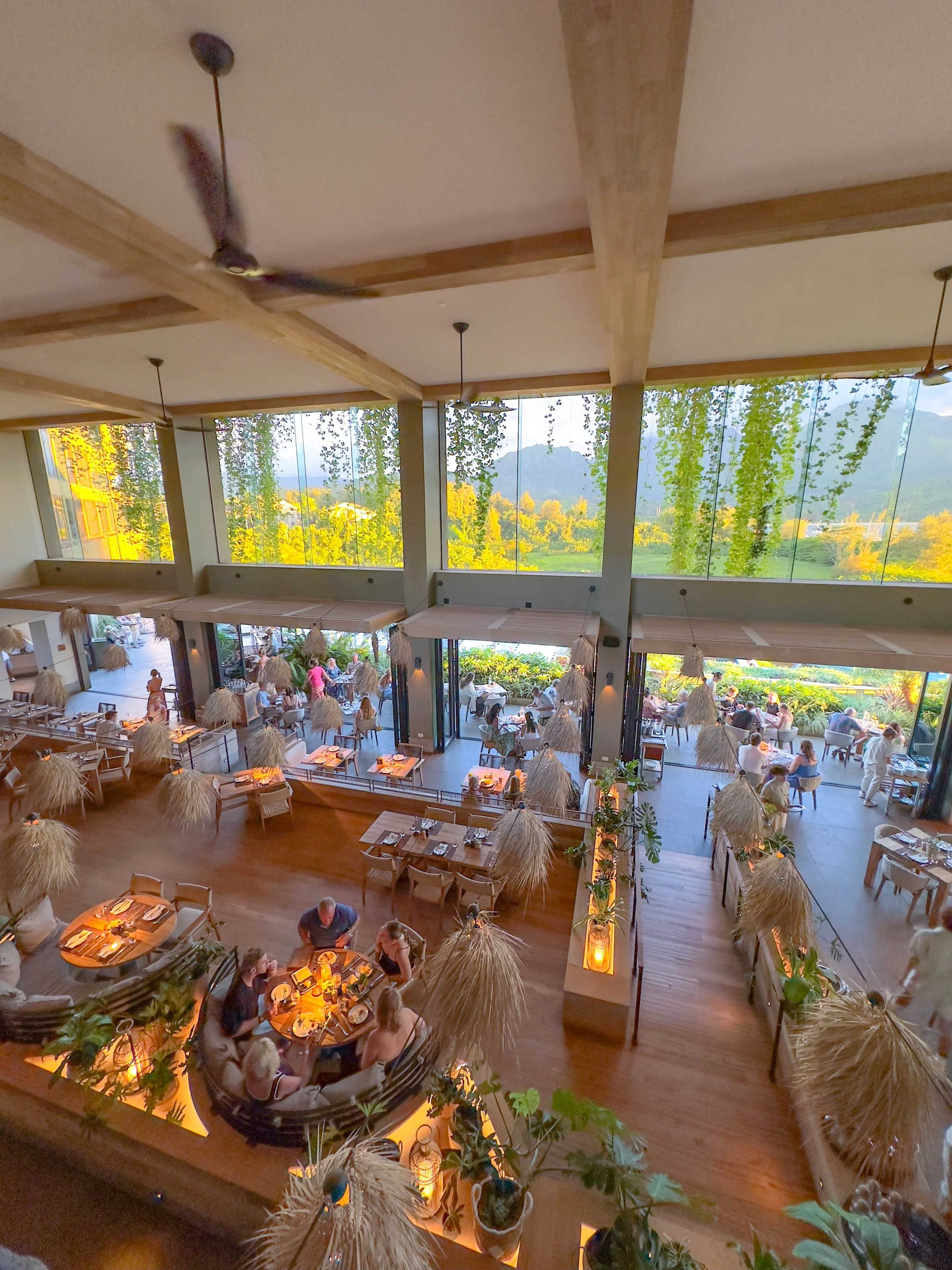 Interior view of the 1Hotel Hanalei Bay. The restaurant is decorated with natural elements like plants and straw umbrellas, and has many diners dining at tables with warm lighting.