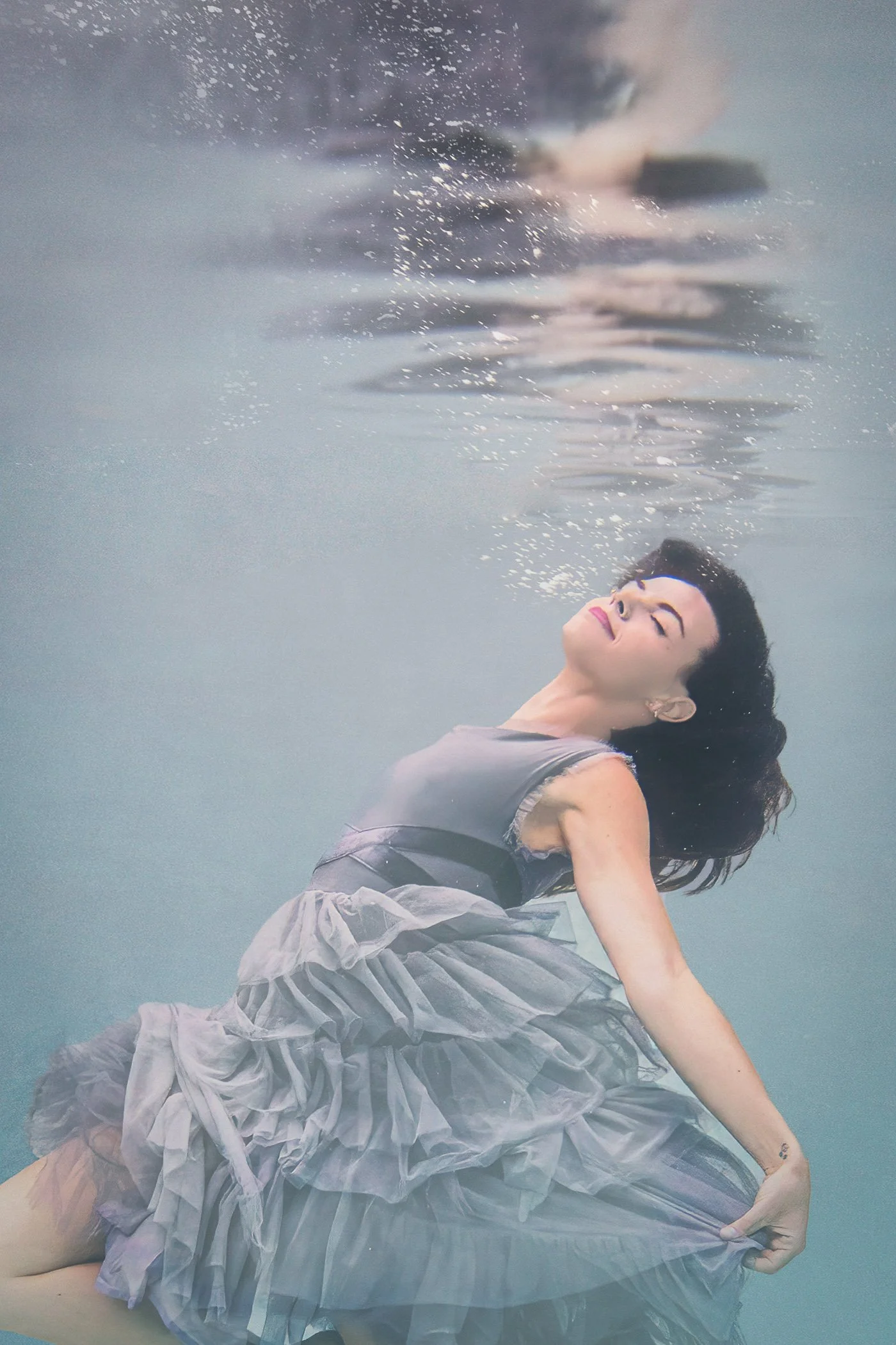 Fine art underwater photograph of a woman suspended beneath the surface wearing a blue frilly dress in pastel blue water.