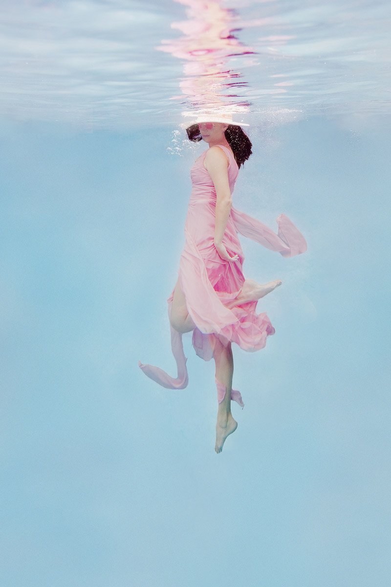 Woman floating underwater in a pink dress wearing sunglasses and a hat, fine art underwater photography