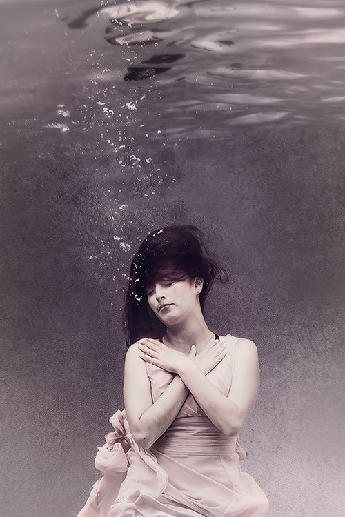 A woman sits quietly underwater in soft light, her form suspended in stillness as fabric drifts gently around her in this fine art photograph.