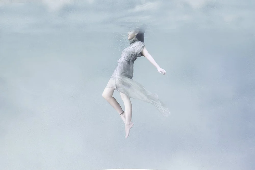 A woman floats just beneath the water’s surface in a sheer dress, her body suspended in soft blue tones in this ethereal underwater fine art photograph.