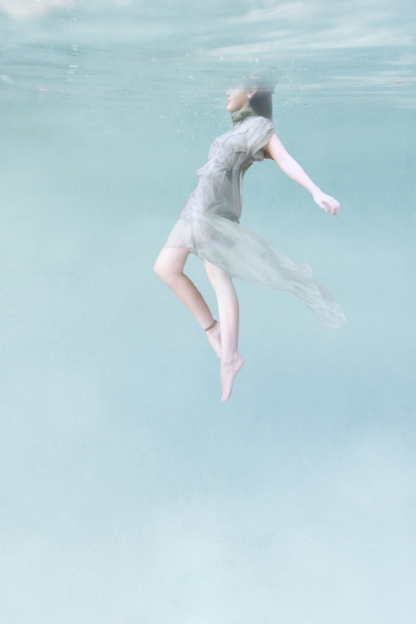 Fine art underwater photograph of a woman in a pastel green-blue pool, wearing a sheer flowing dress with her head partially above the surface.