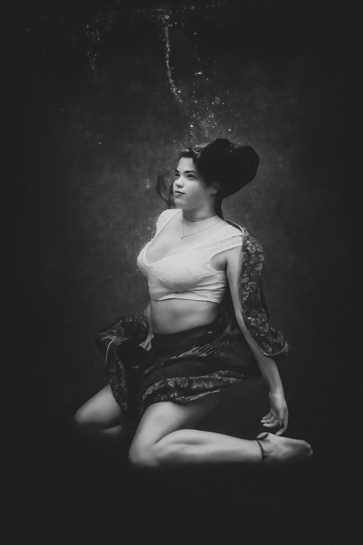 A woman sits underwater in black and white tones, surrounded by flowing fabric and rising bubbles, expressing calm strength and stillness in this fine art photograph.