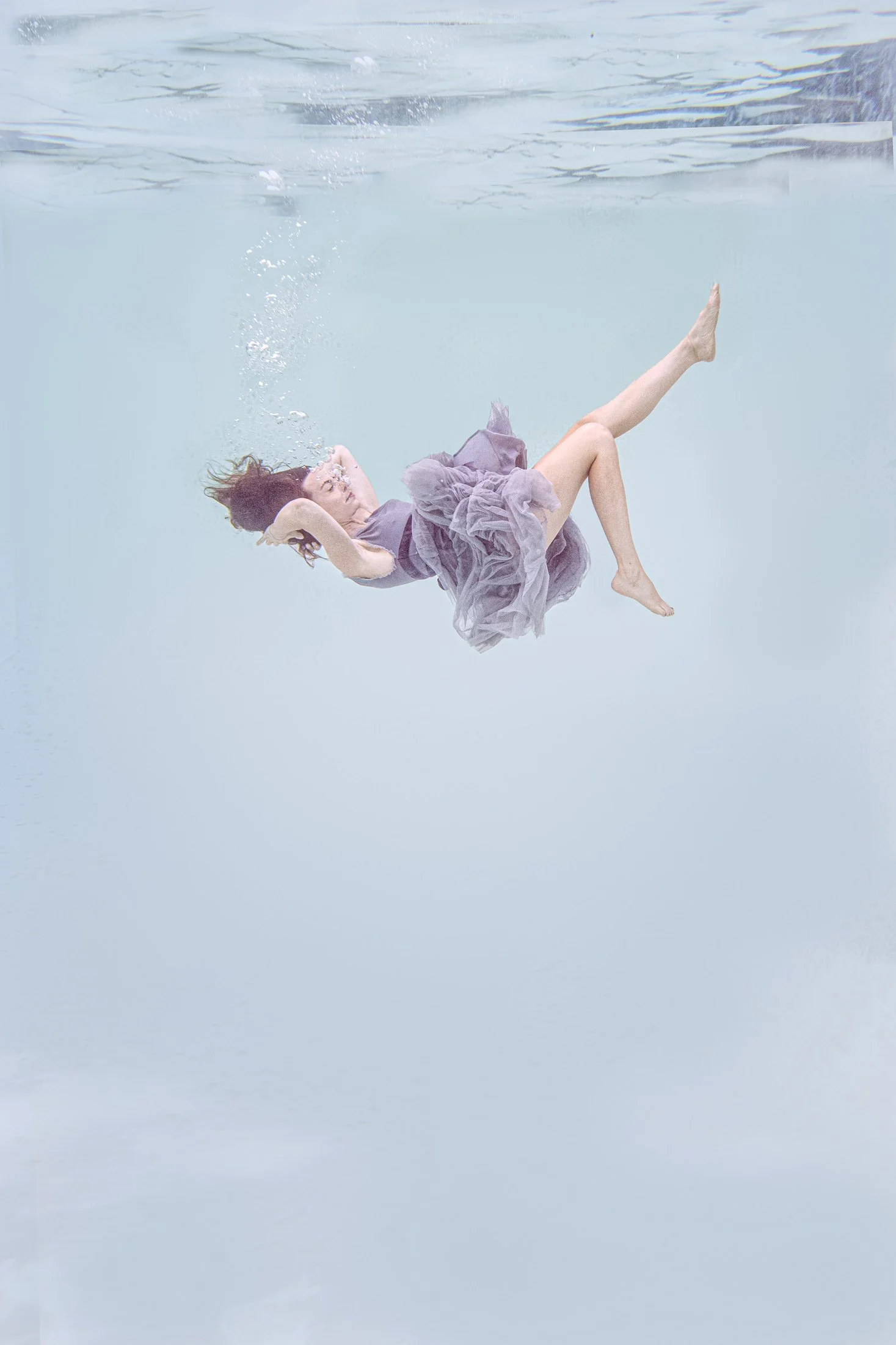 A woman floats beneath the water’s surface in a soft purple dress, bubbles rising as she drifts in pale blue water in this fine art photograph.
