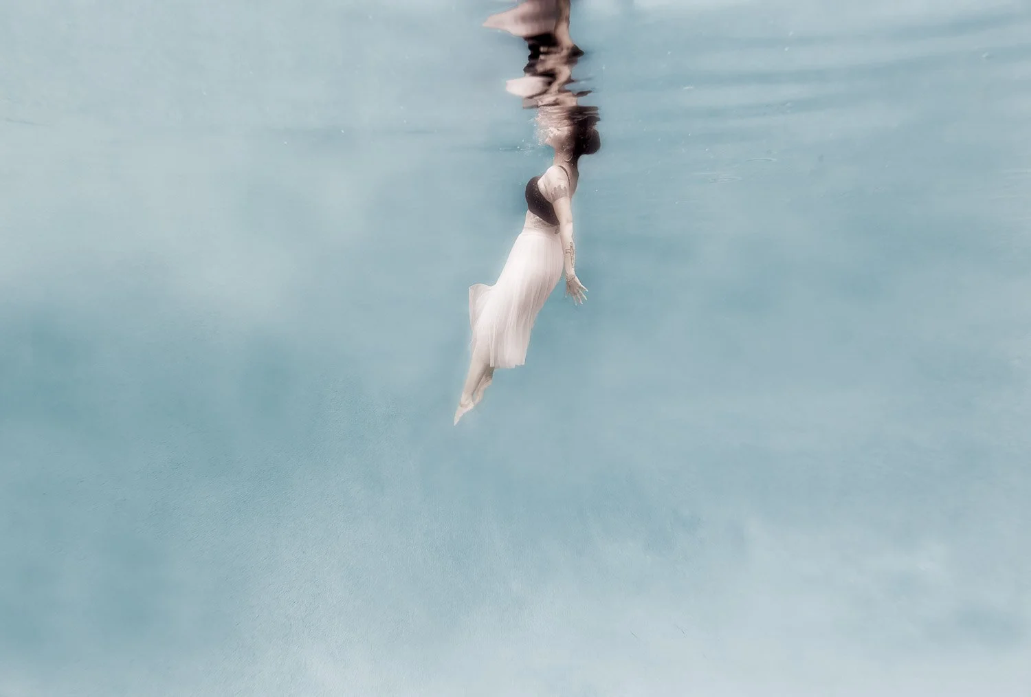 Fine art underwater portrait of a woman floating toward the surface, photographed from below, wearing a pale pink skirt in soft blue water