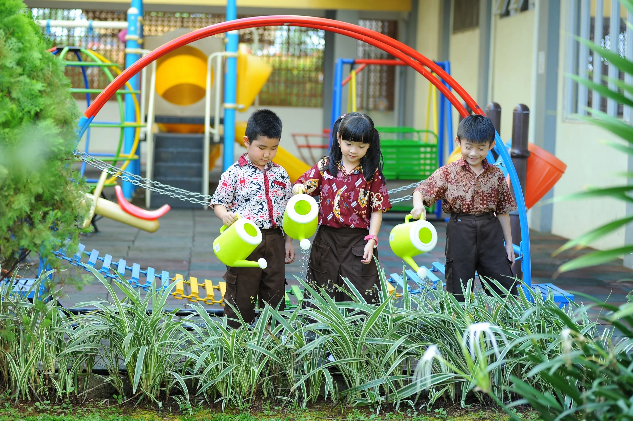 Three children water garden plants with bright green watering cans in a school playground, surrounded by greenery and colorful play equipment that blends outdoor learning with nature.
