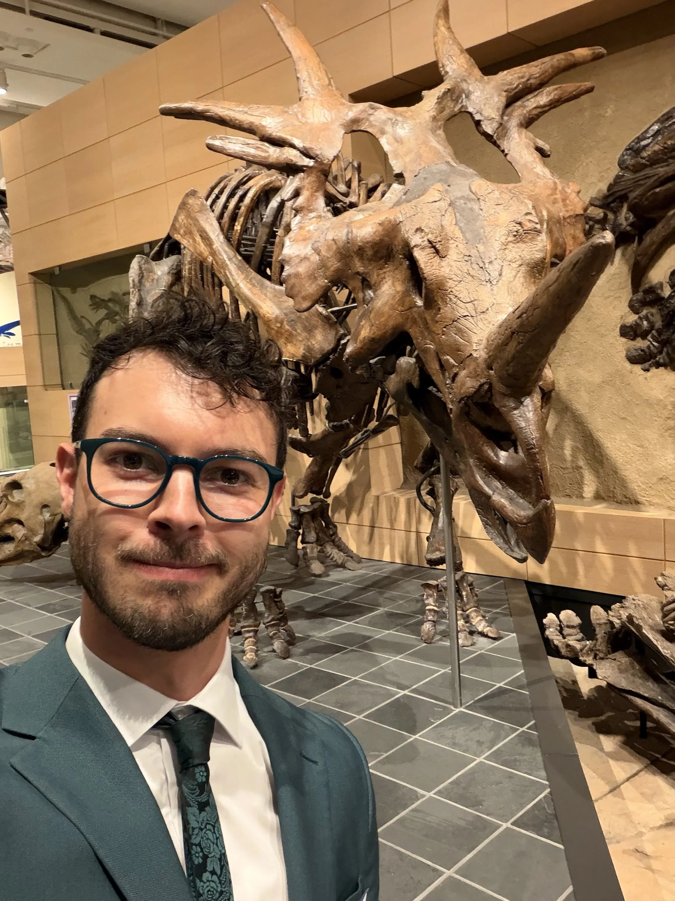 Dr. Owen Wiseman smiling in a teal suit and patterned tie beside a towering Triceratops fossil skeleton at Ottawa's Canadian Museum of Nature, with the dinosaur's enormous skull, three horns and bony frill prominently visible behind him.