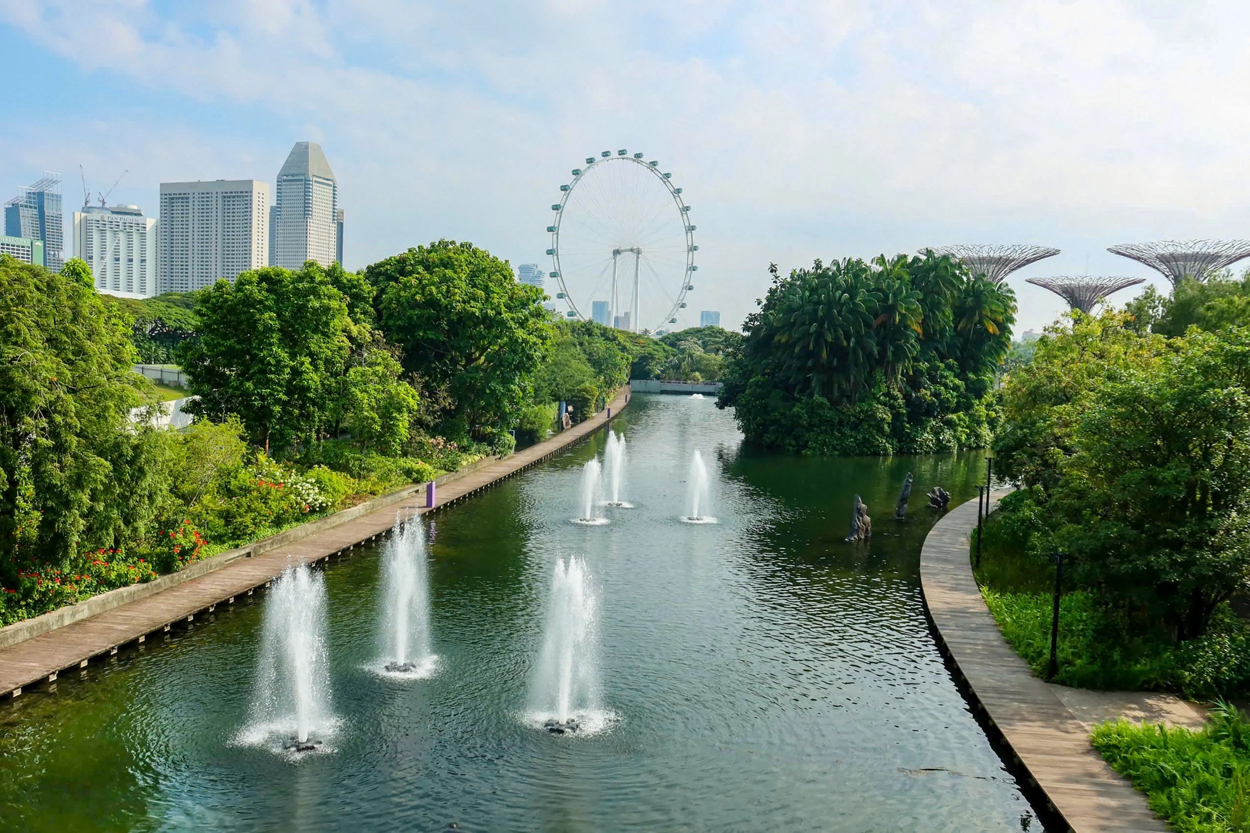 An urban park in Singapore features a tree-lined waterway with fountains, dense tropical vegetation, and walking paths, with the Singapore Flyer and city skyline visible in the background.