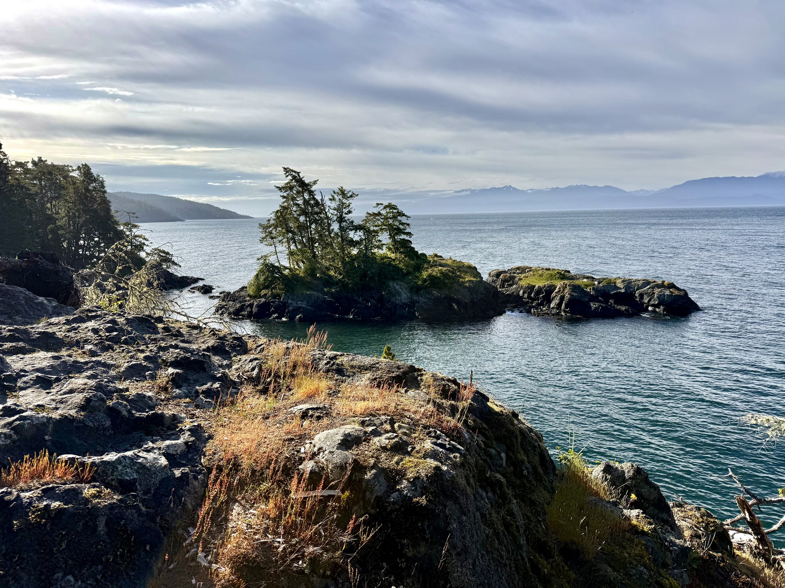 A panoramic view from a rocky coastal bluff in East Sooke Regional Park overlooks calm ocean waters, small forested islets, rugged shoreline, and distant mountains beneath dramatic clouds illuminated by late afternoon sunlight.