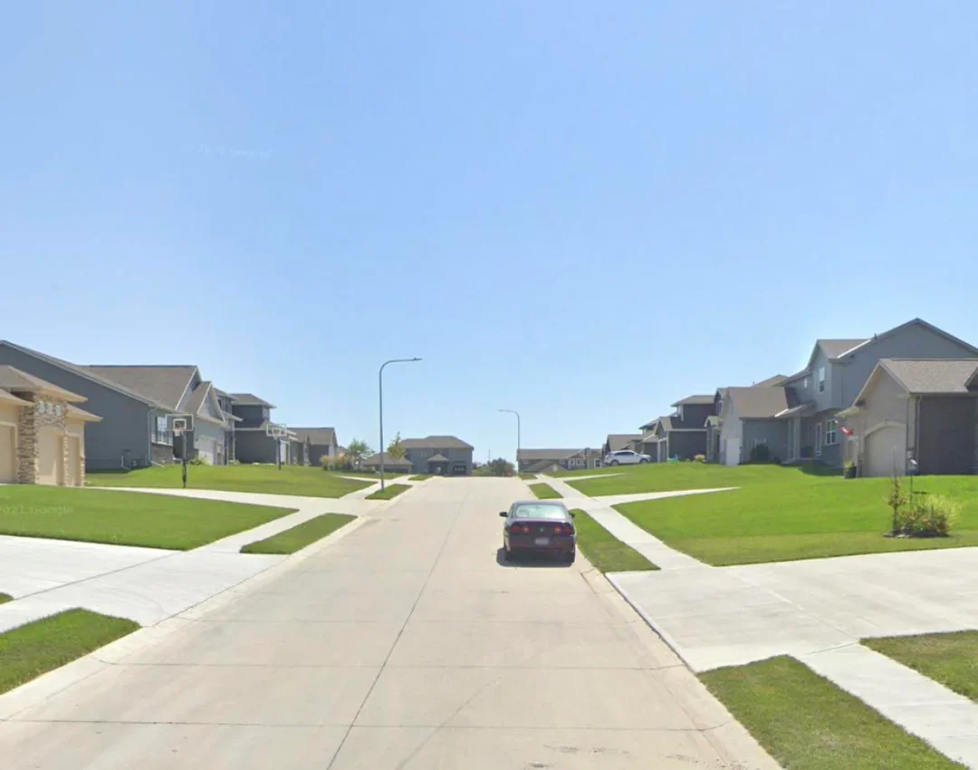 A suburban street with wide roads, large front lawns, and sparse street trees, creating an open neighborhood with limited tree canopy and little shade.