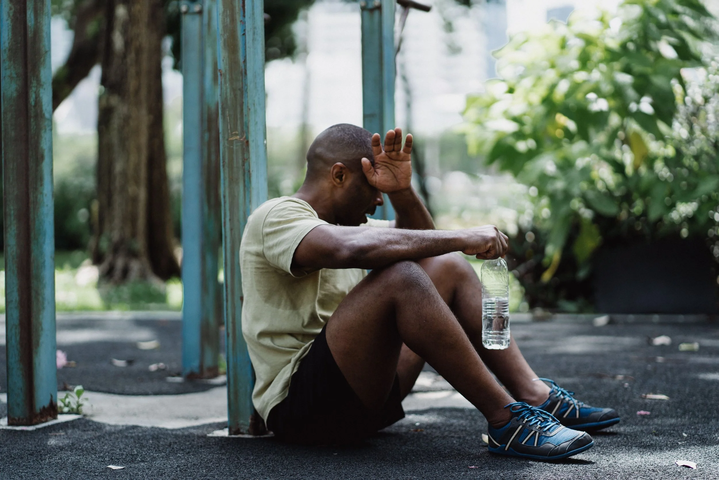 A person sits on the ground outdoors after exercise, shielding their face from the sun while holding a water bottle in a hot, sunny urban environment.