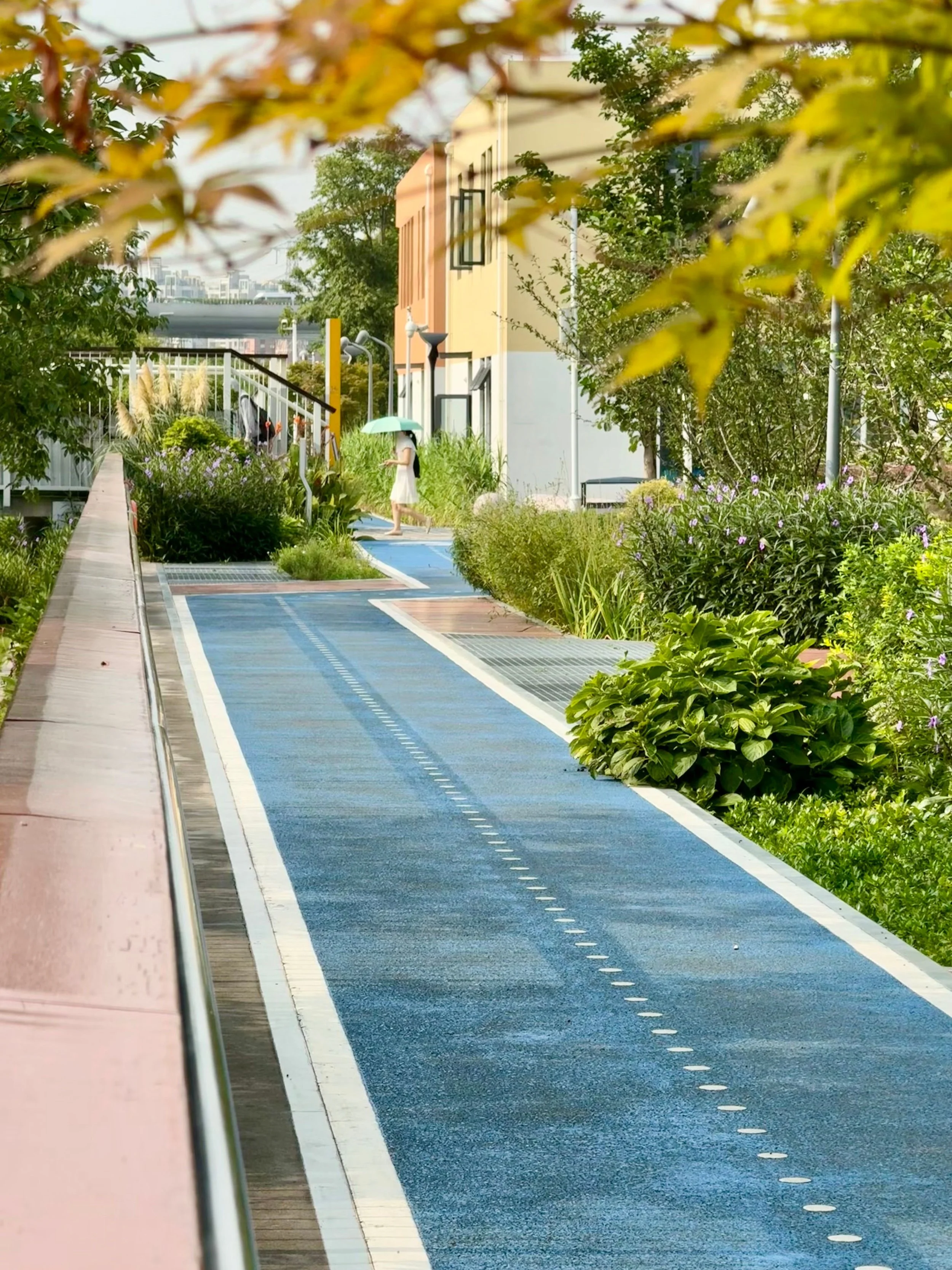 A landscaped urban pathway lined with trees, shrubs, and flowering plants winds between buildings, creating a green, walkable corridor with a person carrying an umbrella in the distance.