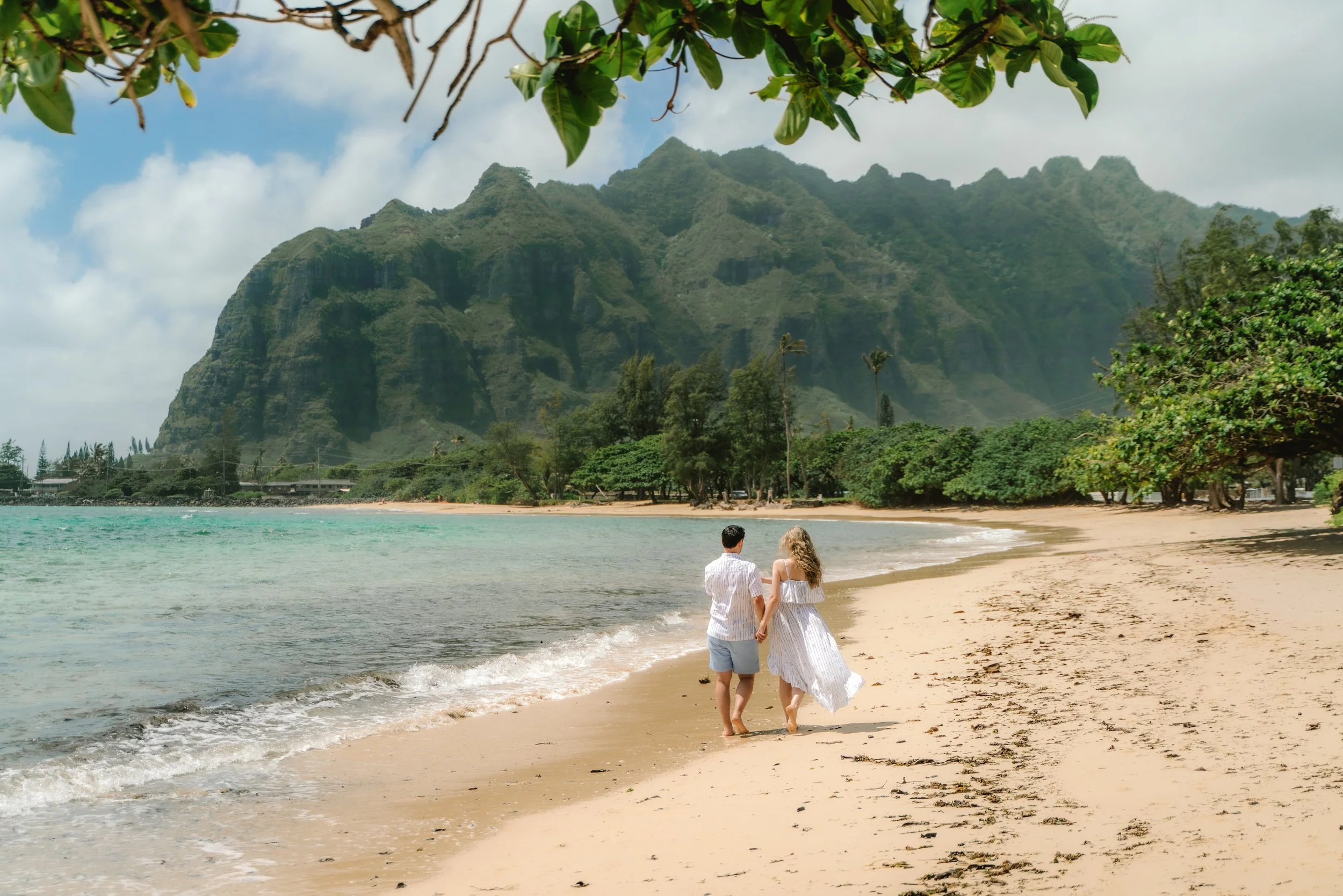 A couple walking hand in hand along a sandy beach near clear turquoise water, with lush green trees and mountainous landscape in the background, under a partly cloudy sky.