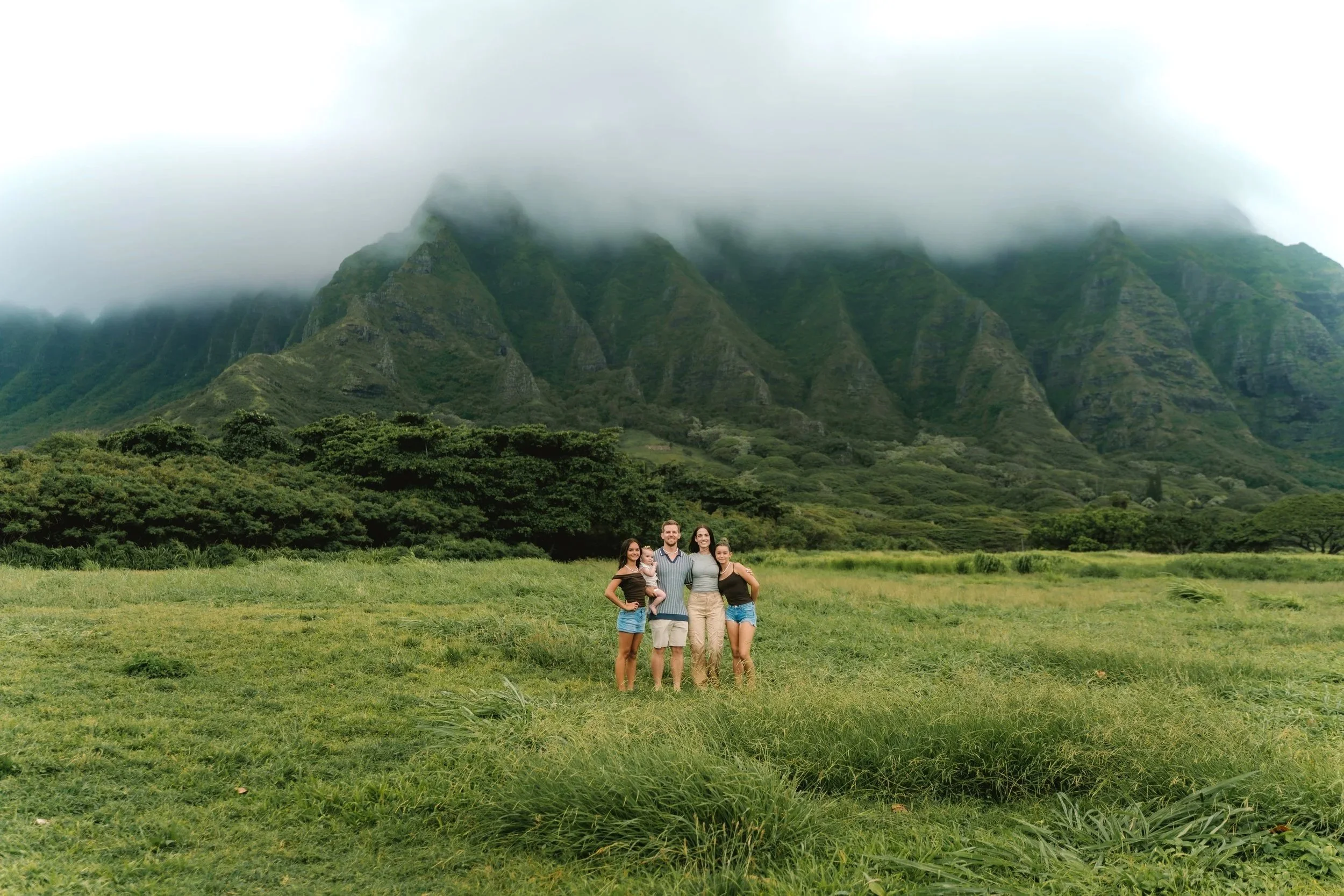 A family of five standing on a grassy field with lush green mountains in the background, partially obscured by clouds.