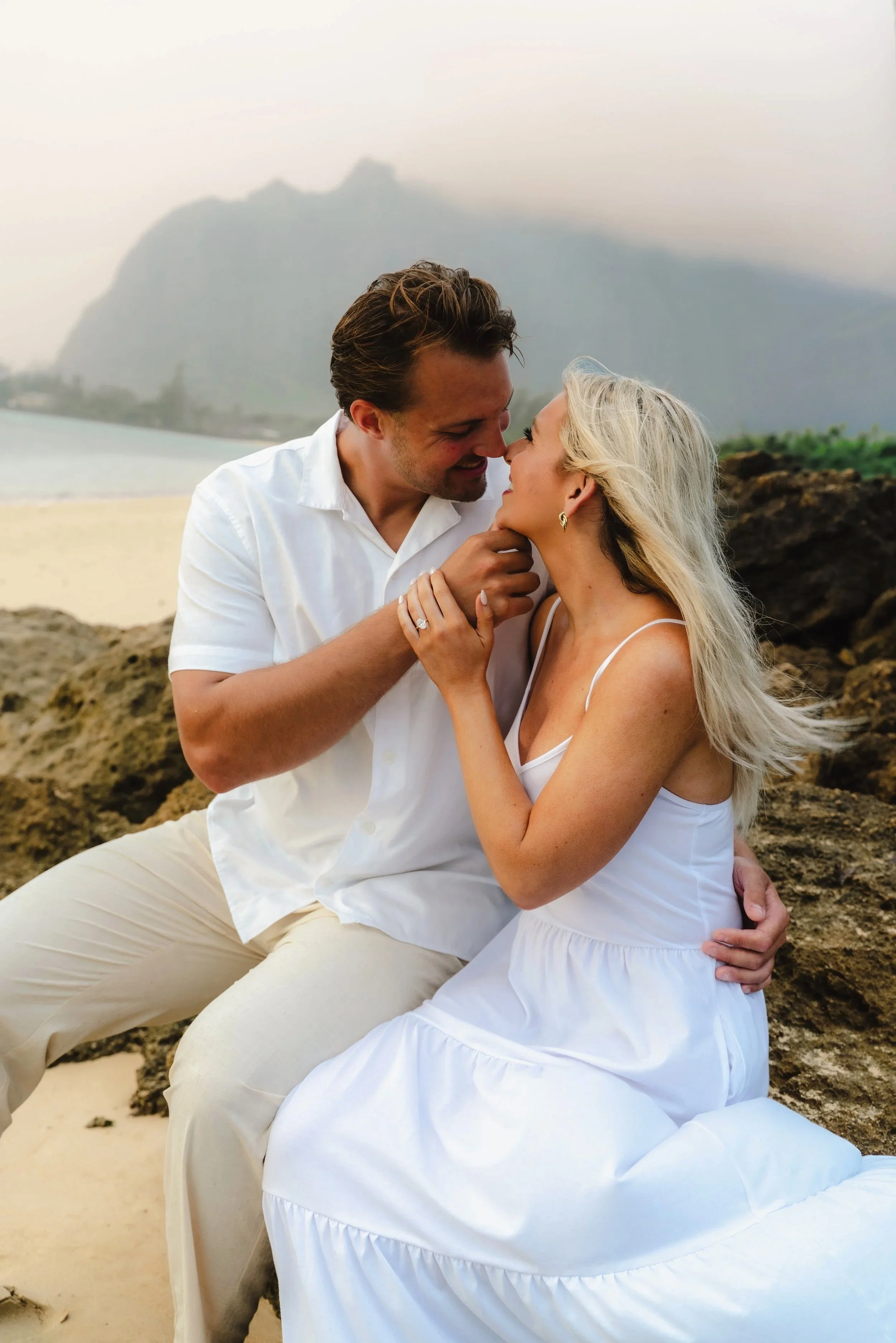 Engaged couple embracing during a sunrise photoshoot at Kaaawa Beach on Oahu with dramatic mountain views and soft morning light.