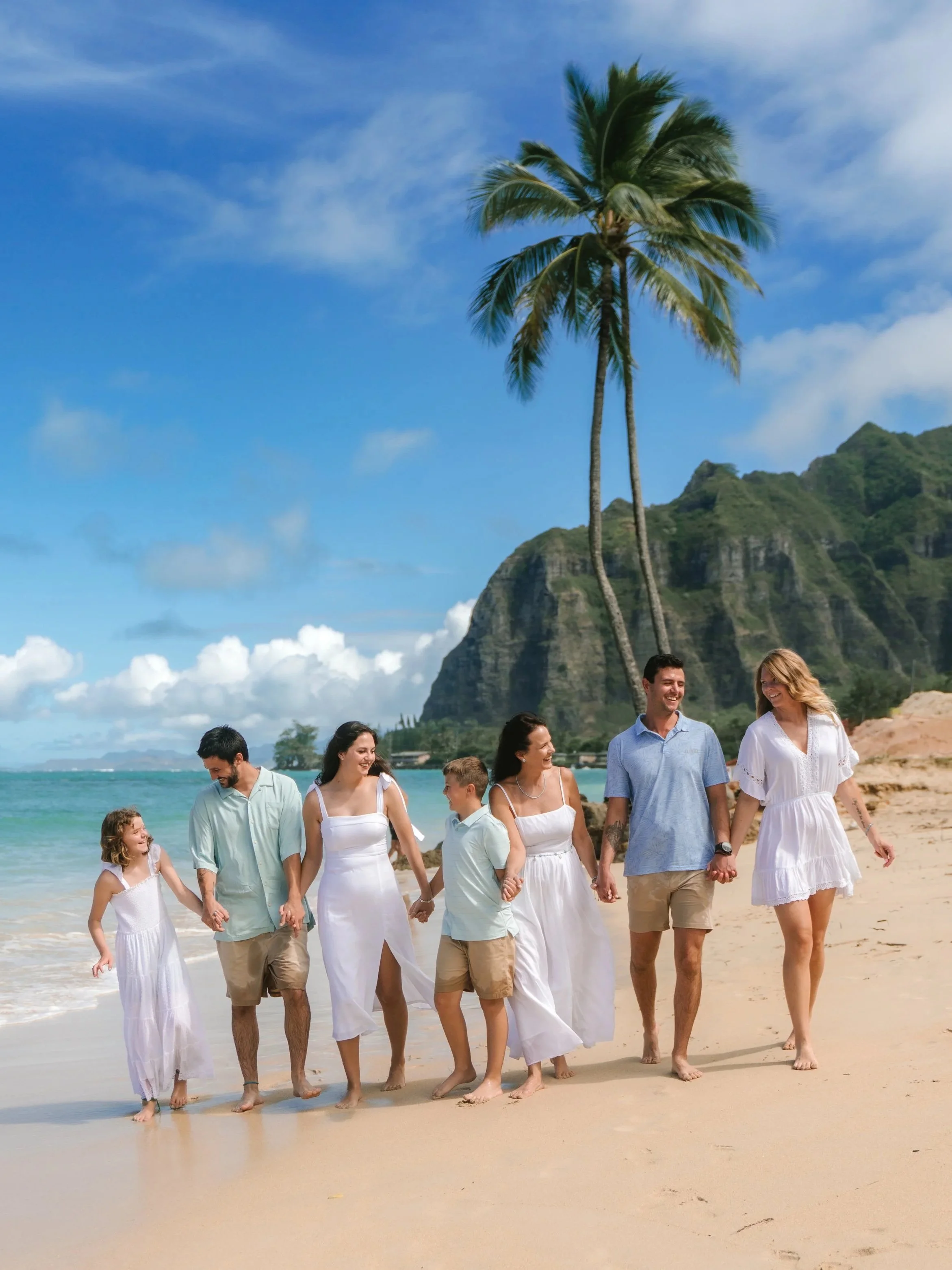Family walking along the beach beneath palm trees with mountain views during a family photography session in Kaaawa, Oahu.