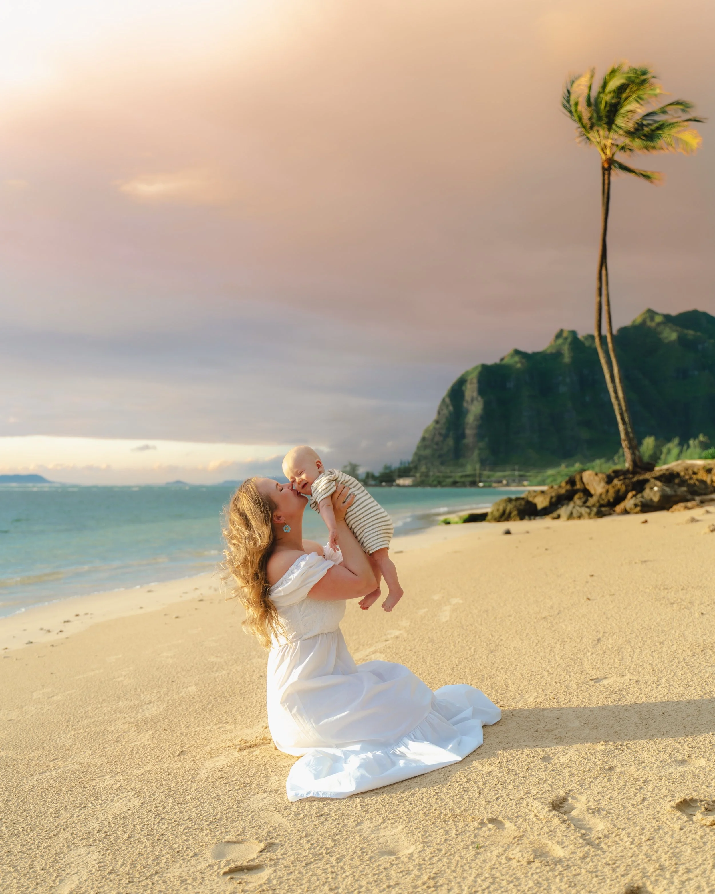 A woman in a white dress kneeling on a sandy beach, holding a smiling baby boy in her arms, with a tall palm tree, rocky hill, and ocean in the background during sunset.