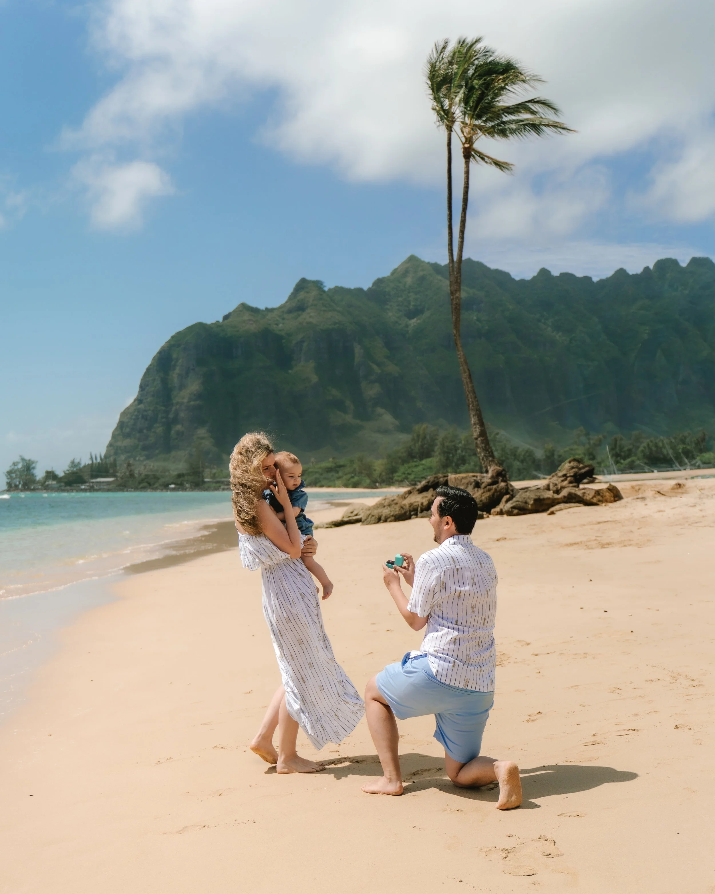 A couple is proposing on a beach with a woman holding a child, a man kneeling with a ring, and family in the background.