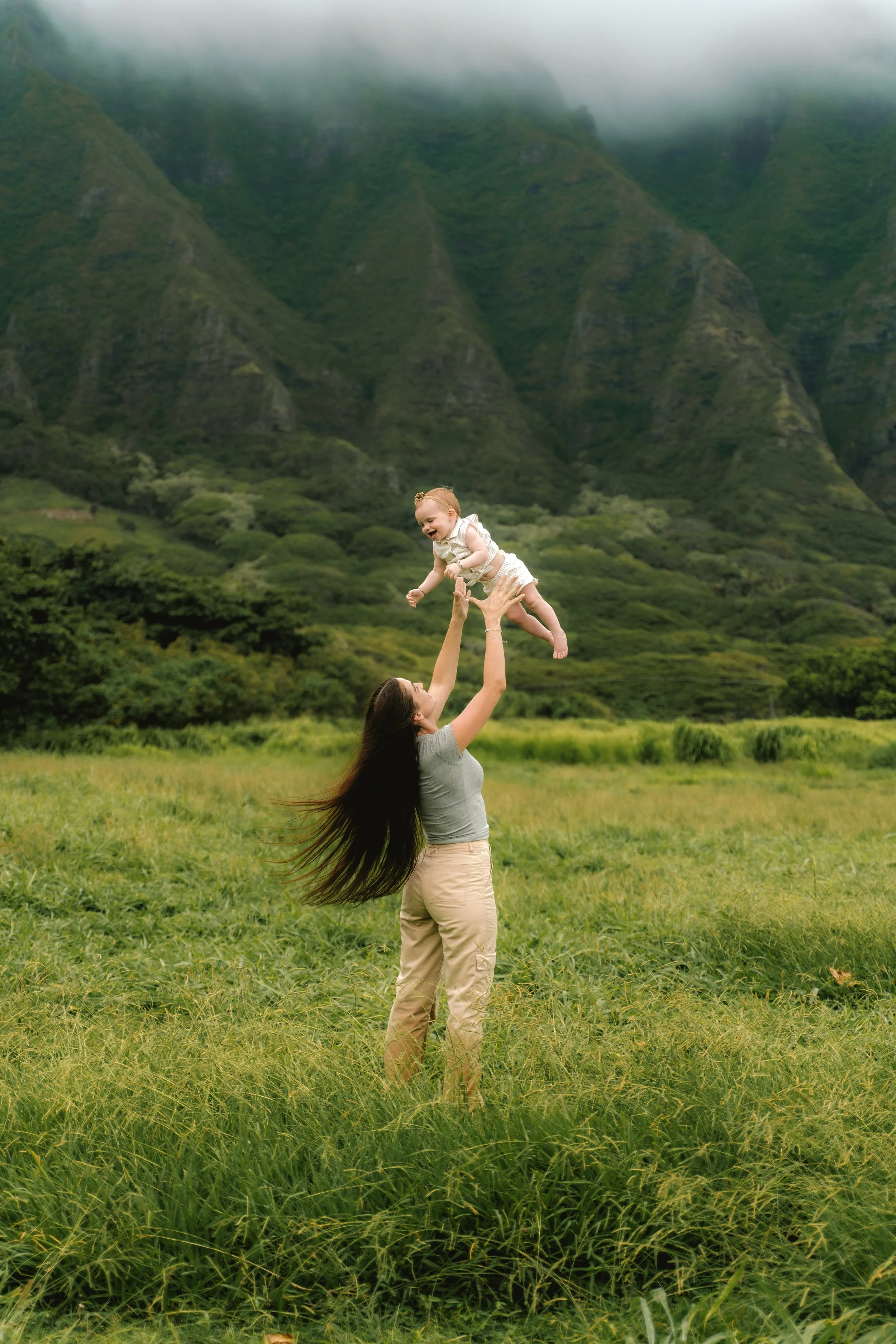 A woman with long dark hair wearing tan pants and a gray shirt lifting a smiling child in white clothing against a lush green mountain landscape with fog.