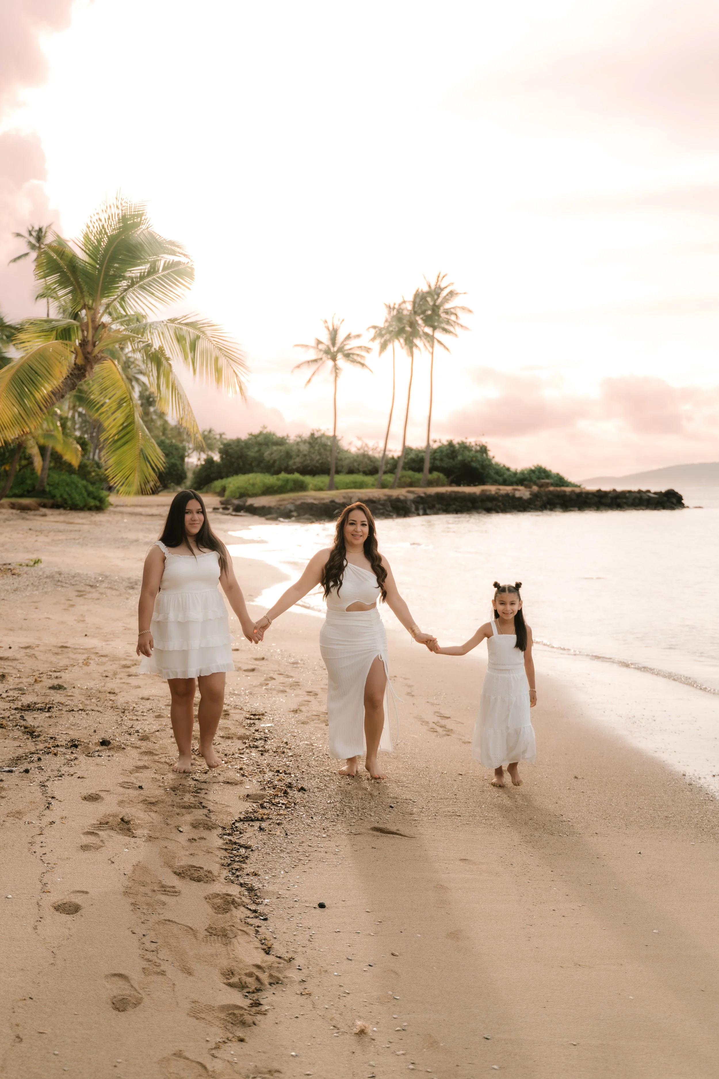 Three women walking hand in hand along the beach with palm trees, rocky shoreline, and sunset sky in the background.