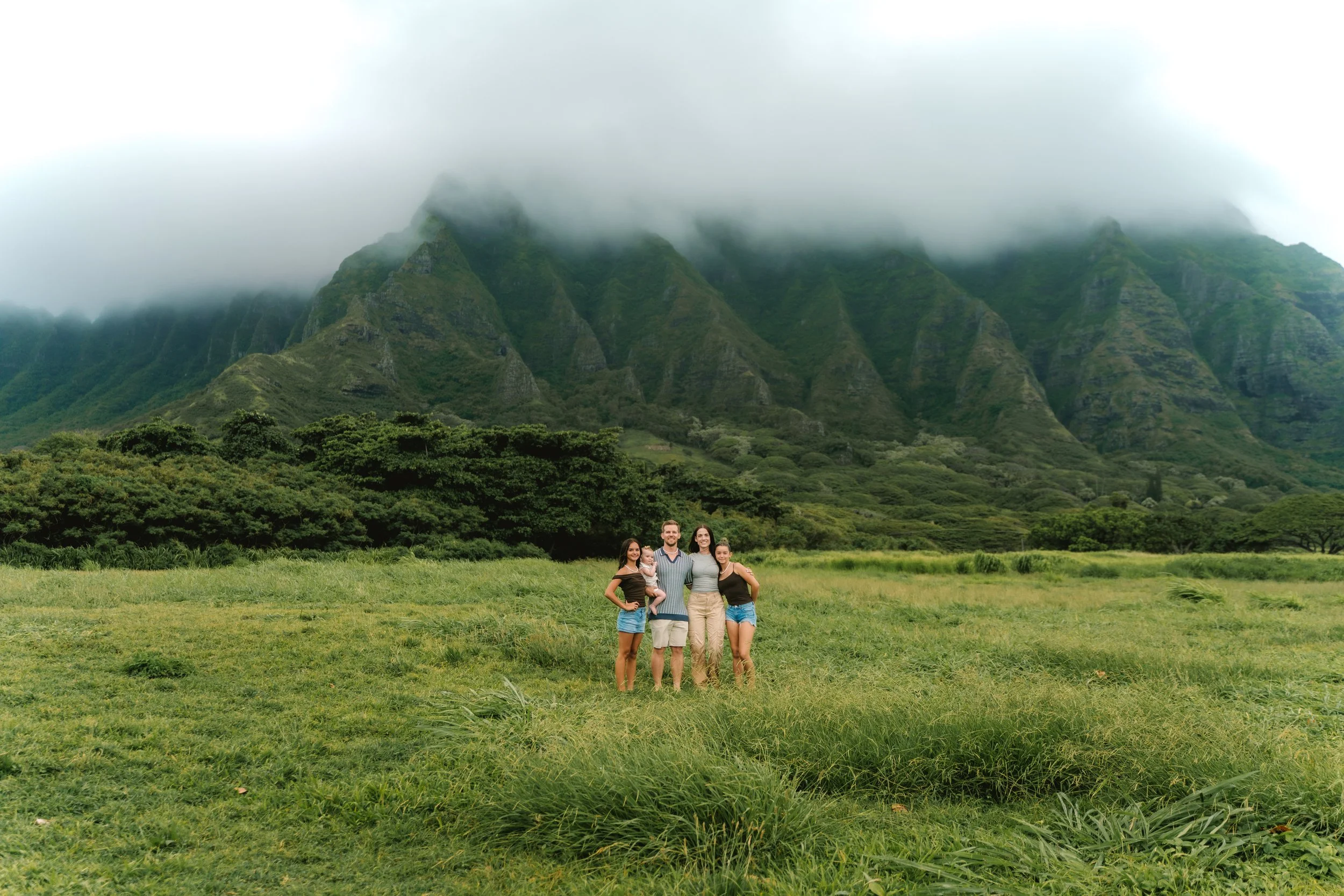 A family of five standing in a grassy field with green mountains and fog in the background.