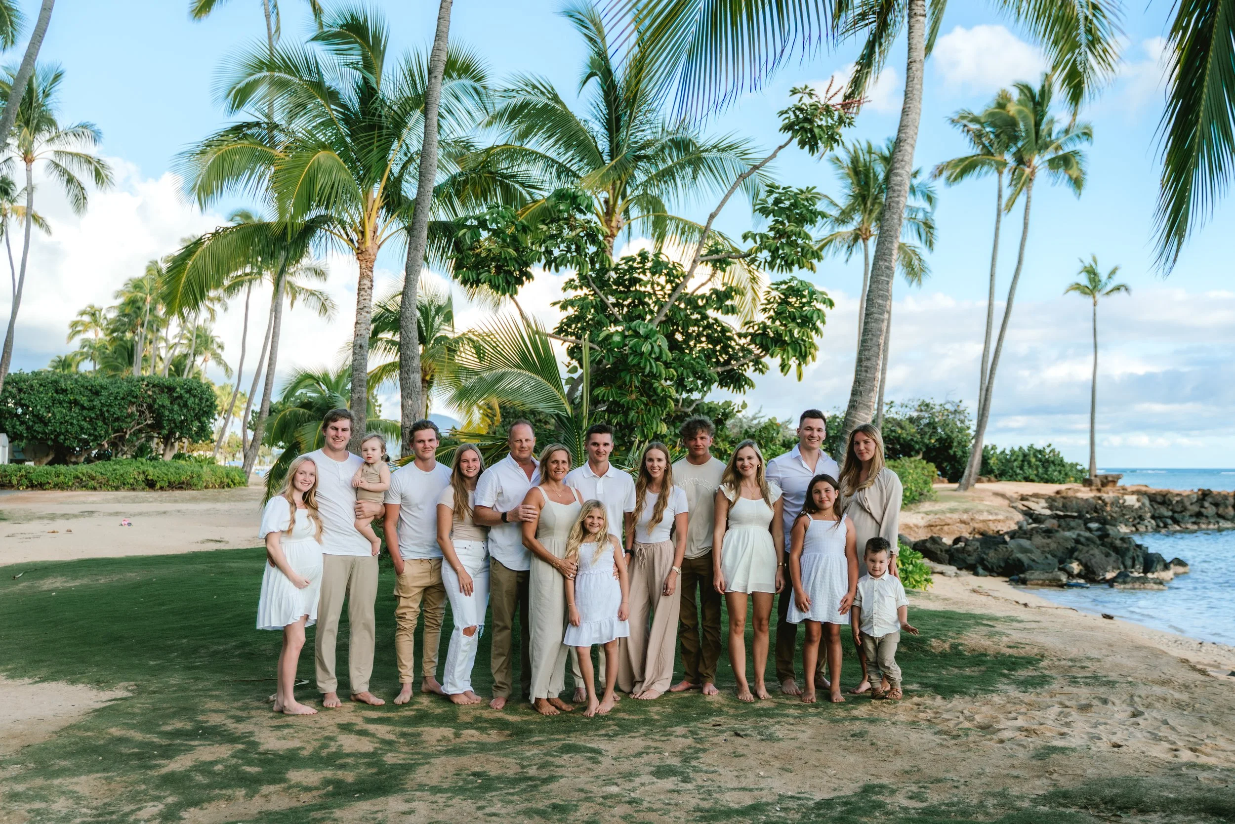 Group of people wearing white and beige clothing standing on a beach with palm trees, sand, and the ocean in the background.