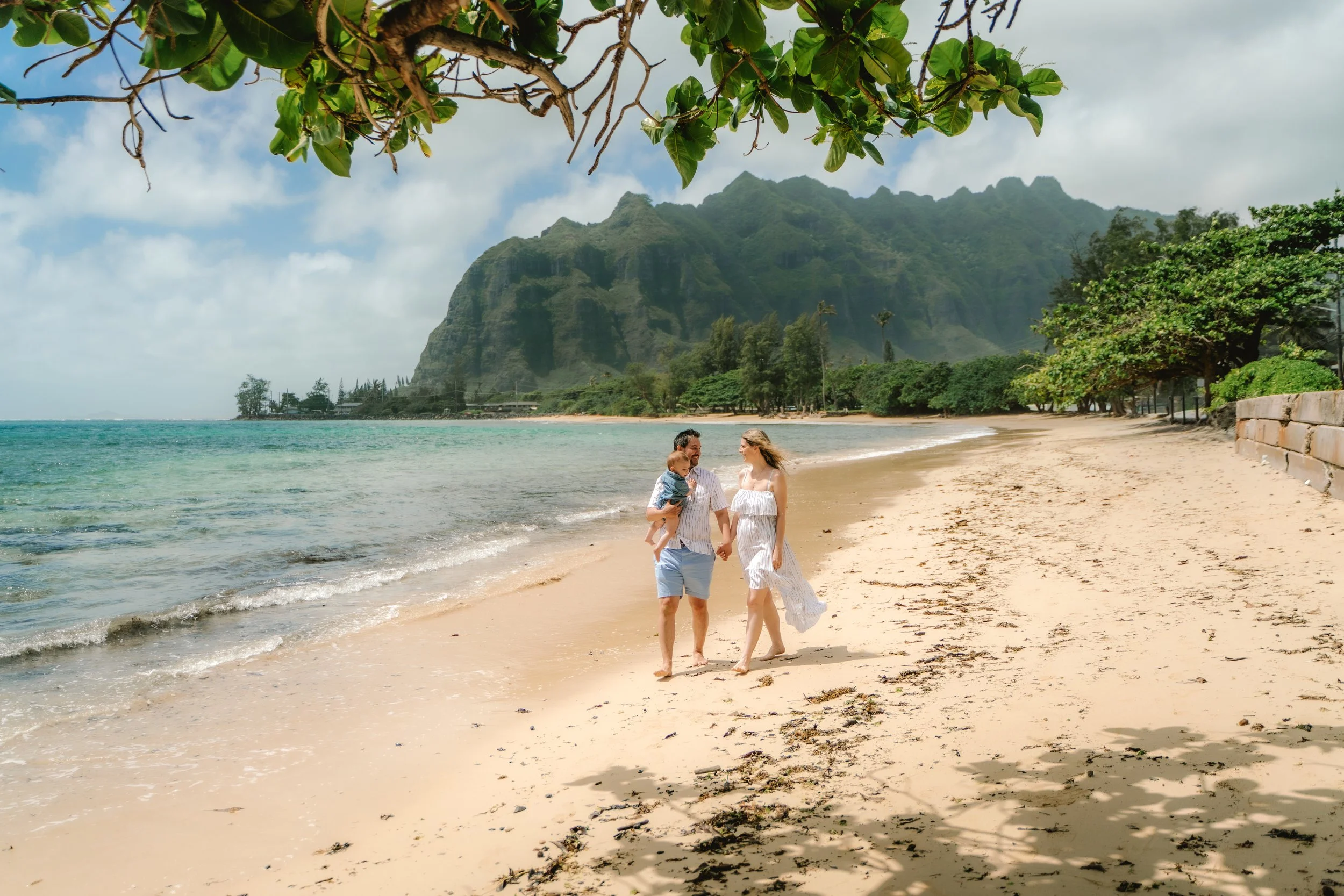 A family walking along a sandy beach with mountains and lush greenery in the background, under a partly cloudy sky.