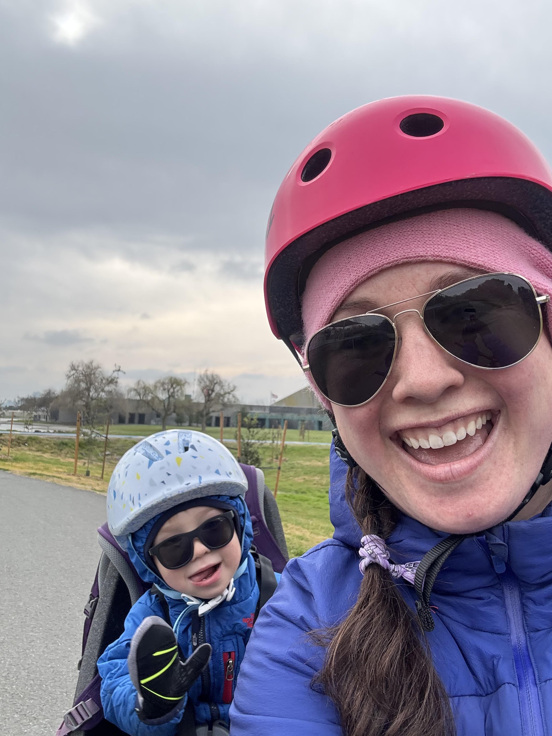 A woman and a young child outdoors wearing helmets and sunglasses, with the woman taking a selfie, on a cloudy day with a park in the background.