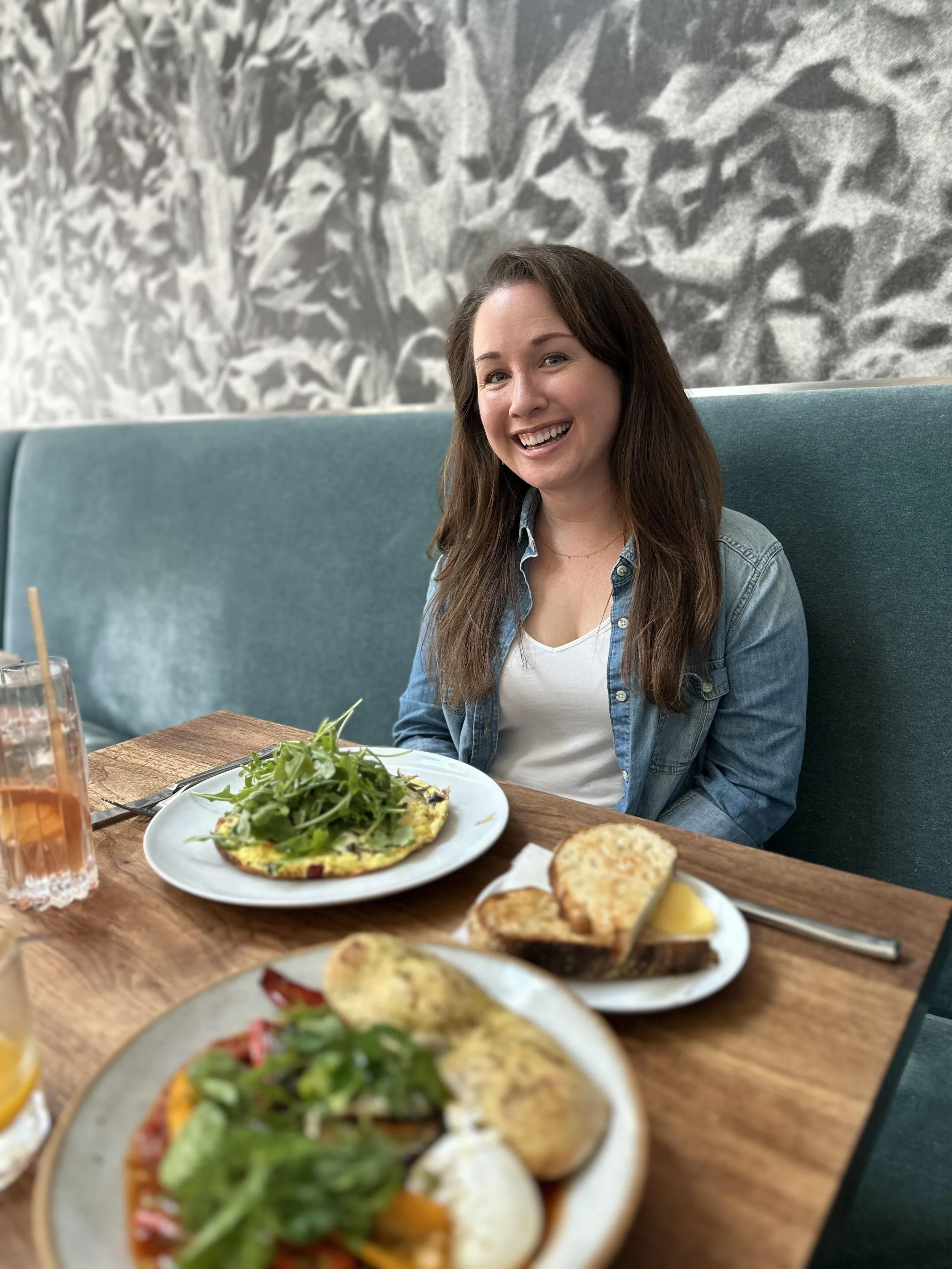 A woman sitting at a restaurant table with plates of food, smiling at the camera. The table has a salad, bread, and drinks.